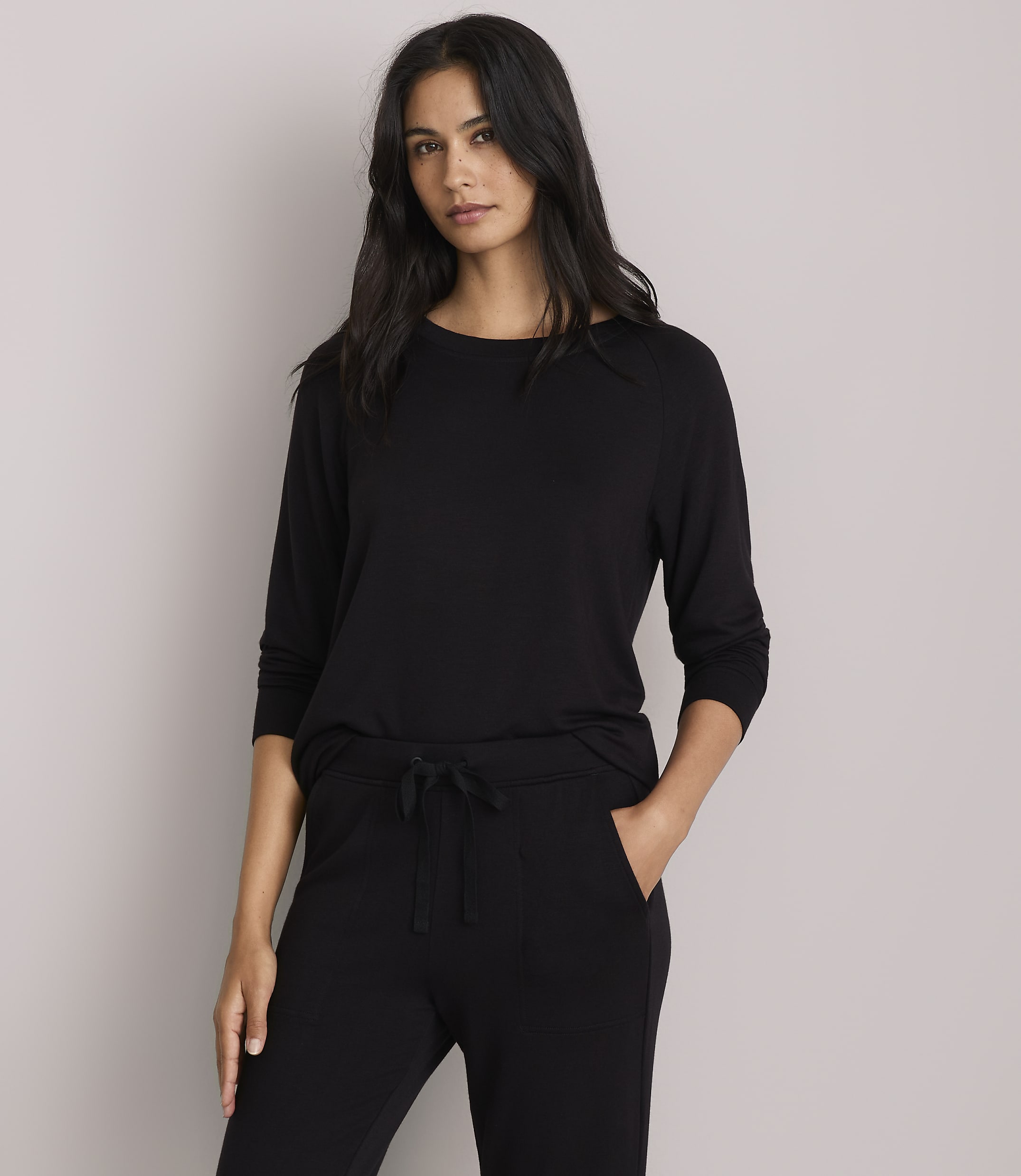 A woman in a black long-sleeved shirt and black pants stands against a plain gray background.