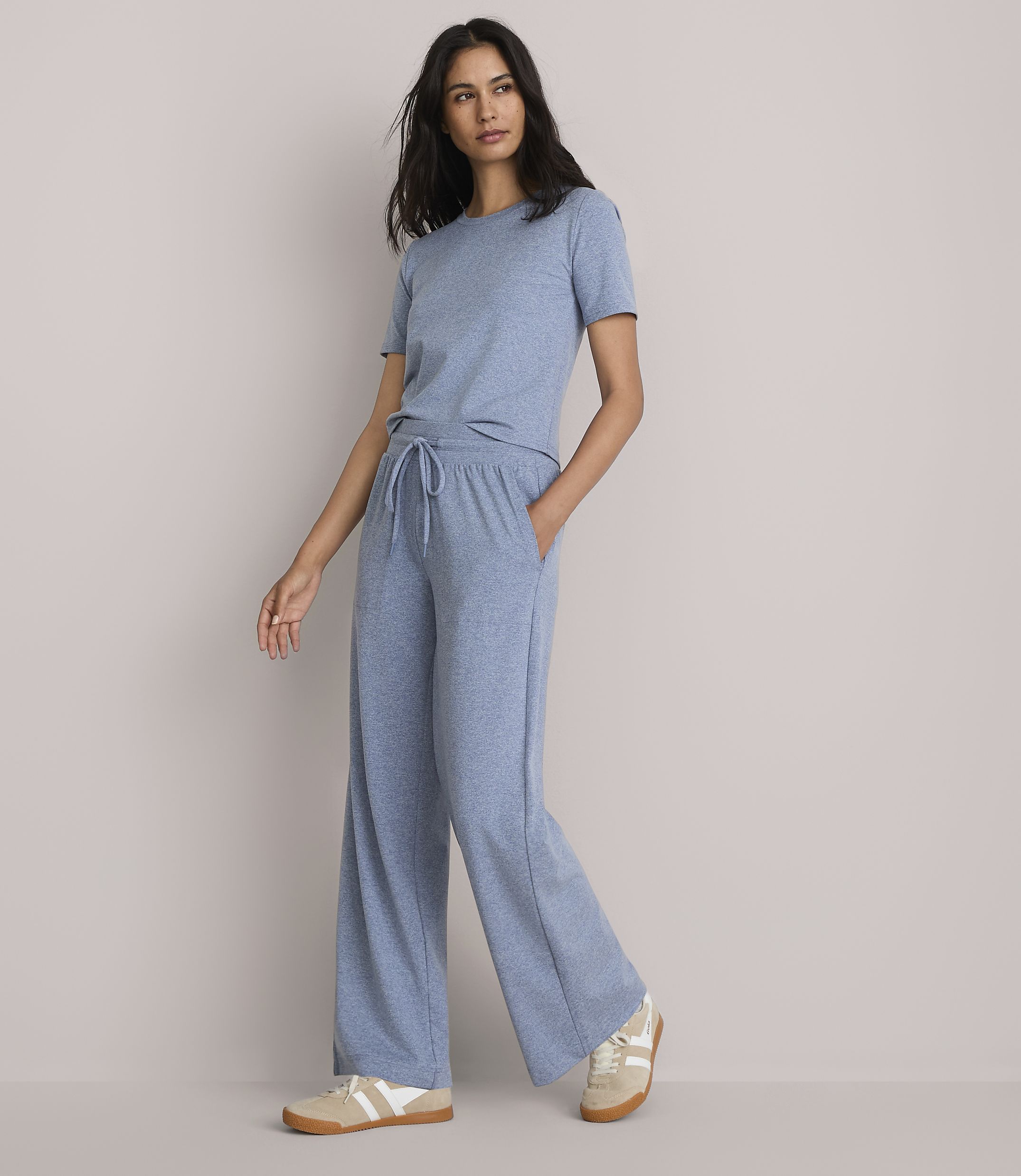 A woman in a blue t-shirt and wide-leg pants stands against a plain, light gray background.