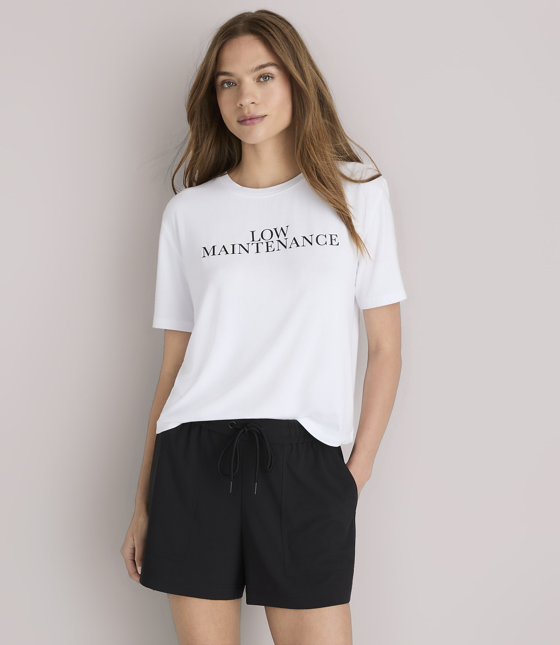 A woman in a white "LOW MAINTENANCE" t-shirt and black baseball cap poses against a plain white background.