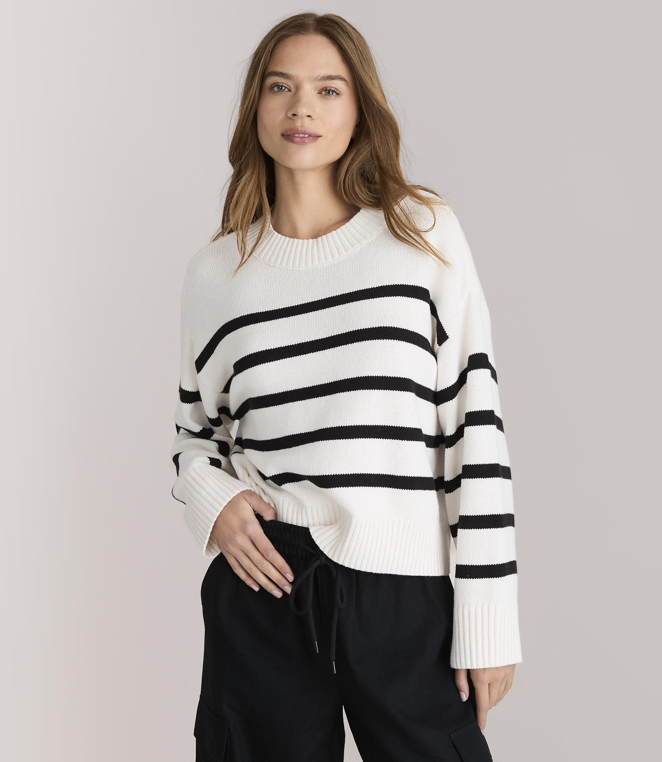 A young woman in a striped sweater and black pants sits cross-legged against a plain white background.