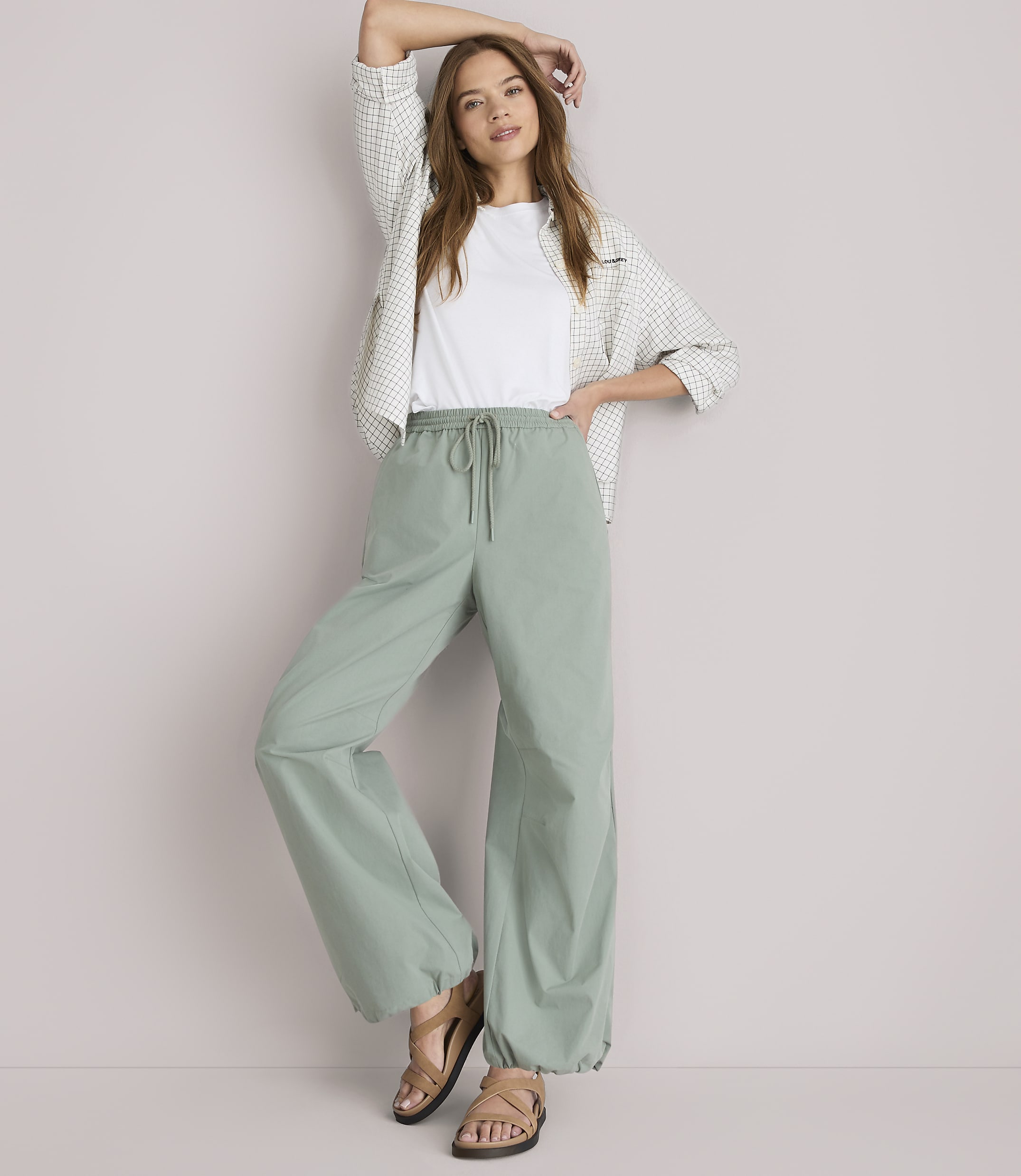 A woman in a white t-shirt, patterned shirt, and loose green pants stands against a plain white background.