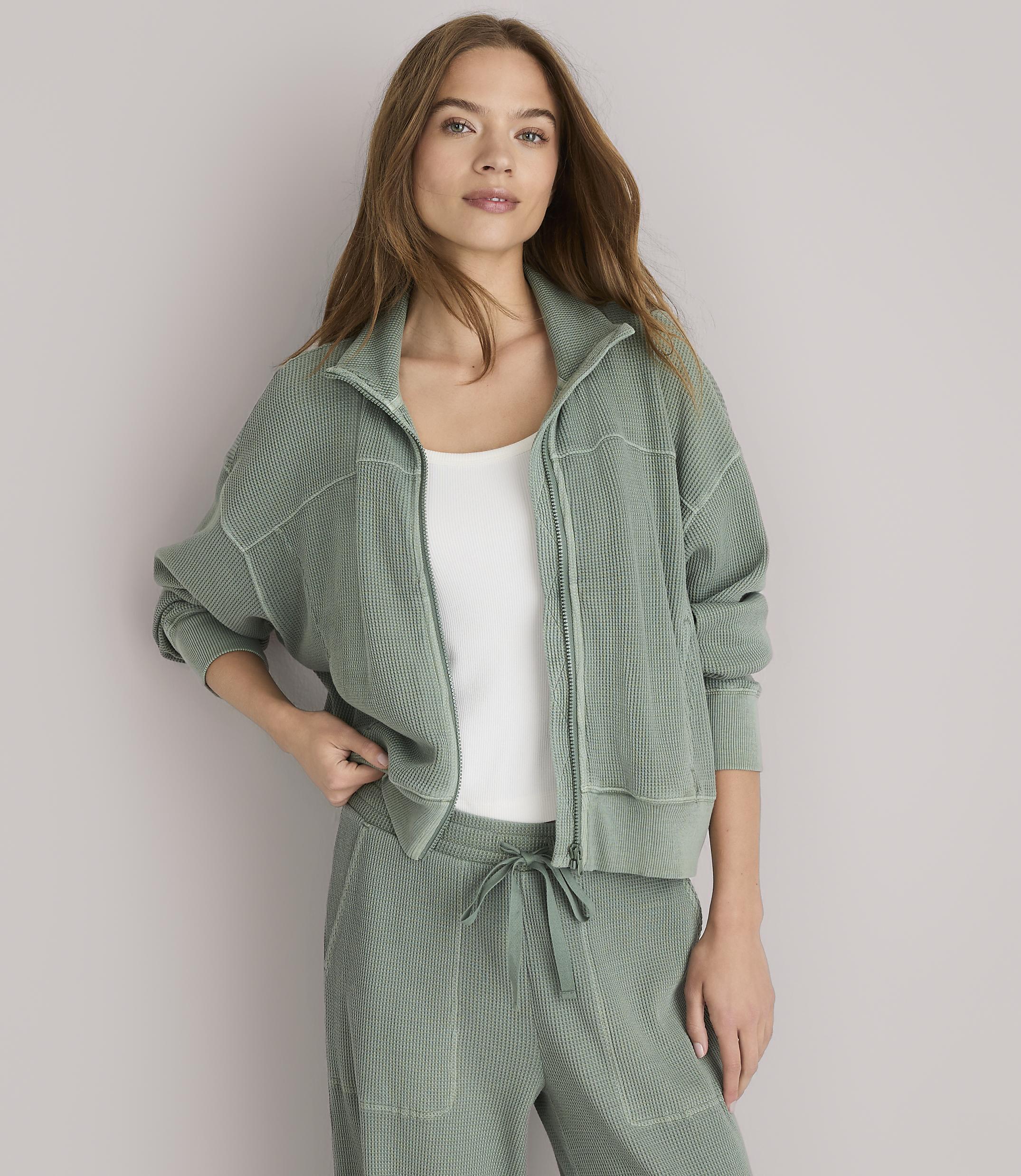 A woman in a green zip-up jacket and matching pants stands against a plain white background.