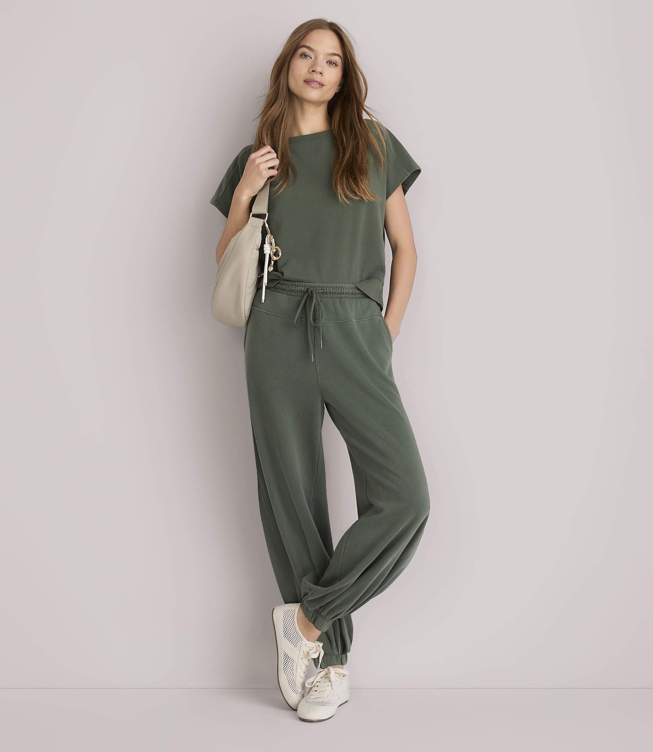 A woman in a green loungewear set and sneakers stands with a cream-colored bag slung over her shoulder against a plain white background.