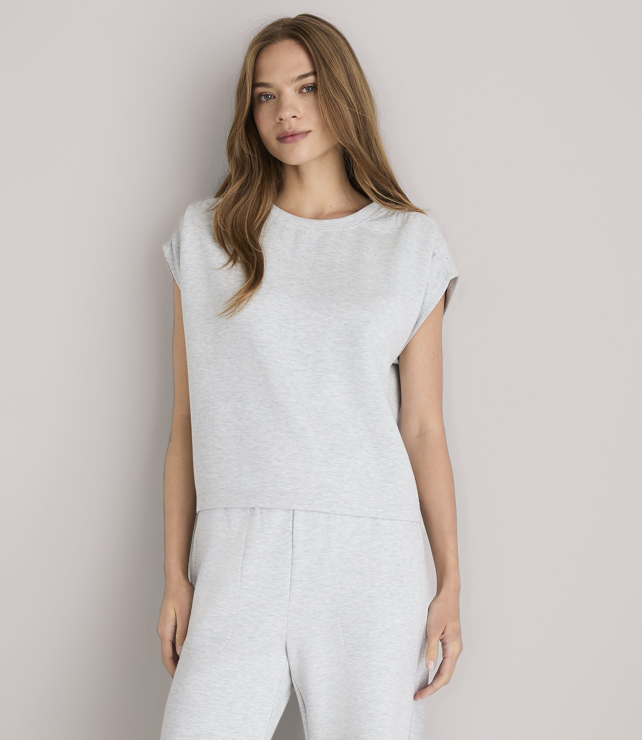 A young woman in a grey loungewear set and white sandals poses against a plain grey background.