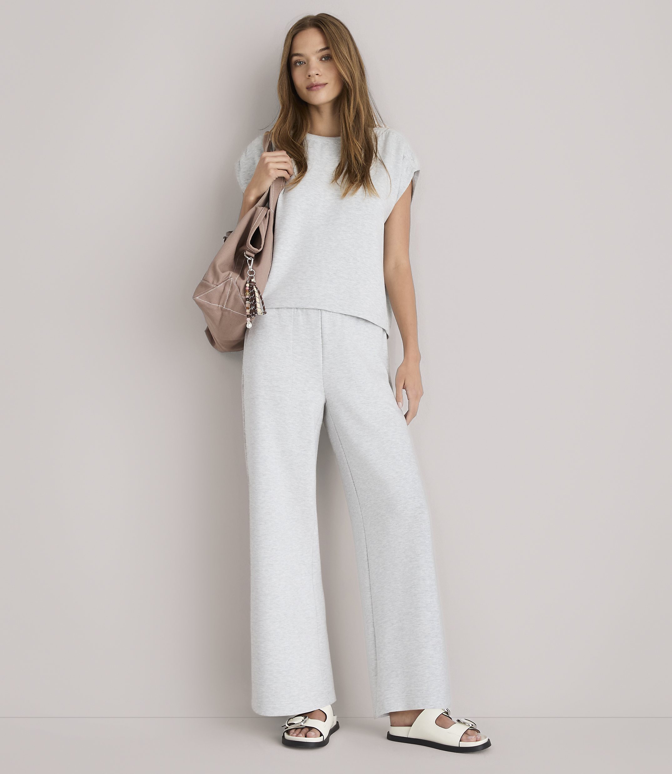 A young woman in a grey loungewear set and white sandals stands against a plain white background, with a pink backpack slung over her shoulder.
