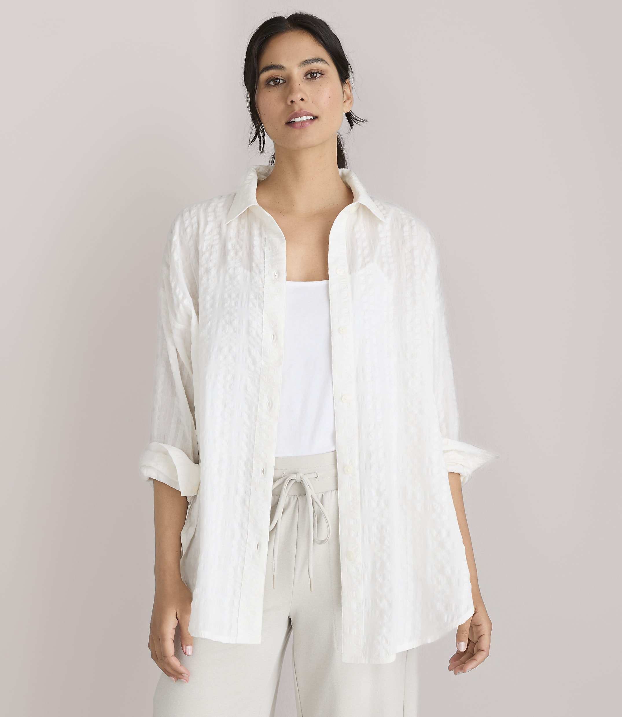 A woman in a white textured shirt and light-colored pants stands against a plain white background.