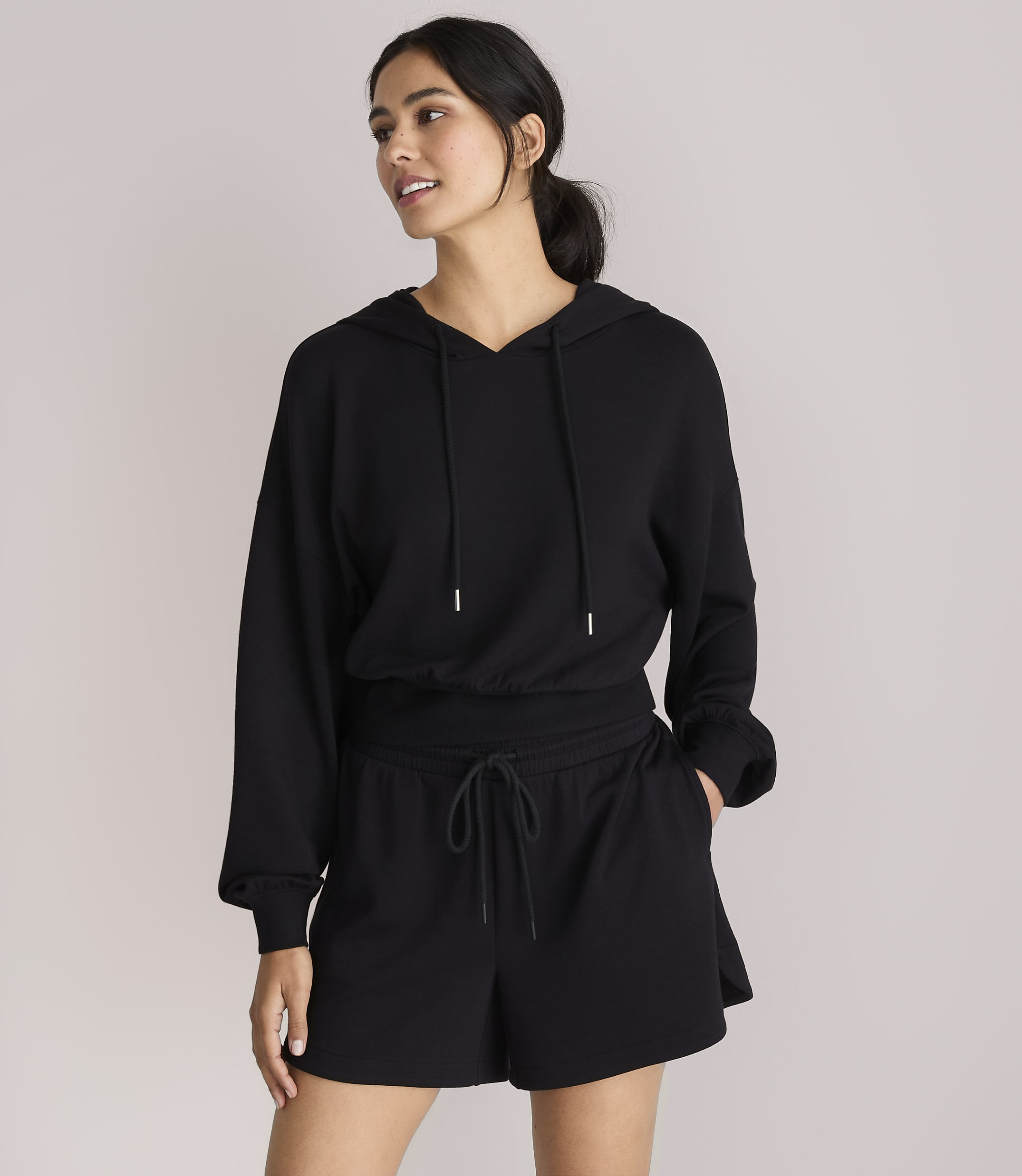 A woman in a black hoodie and shorts stands against a plain white background.