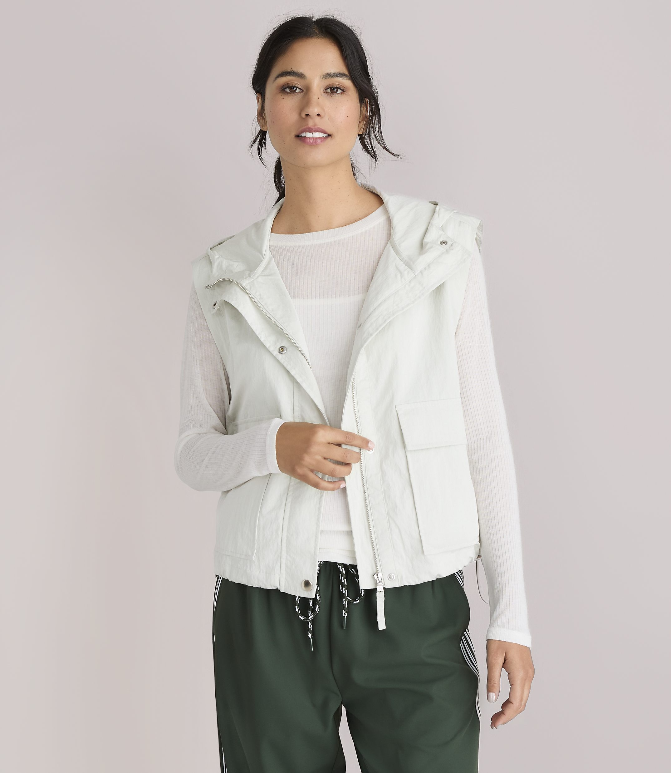 A woman in a light-colored hooded vest and white shirt looks to the side against a plain white background.