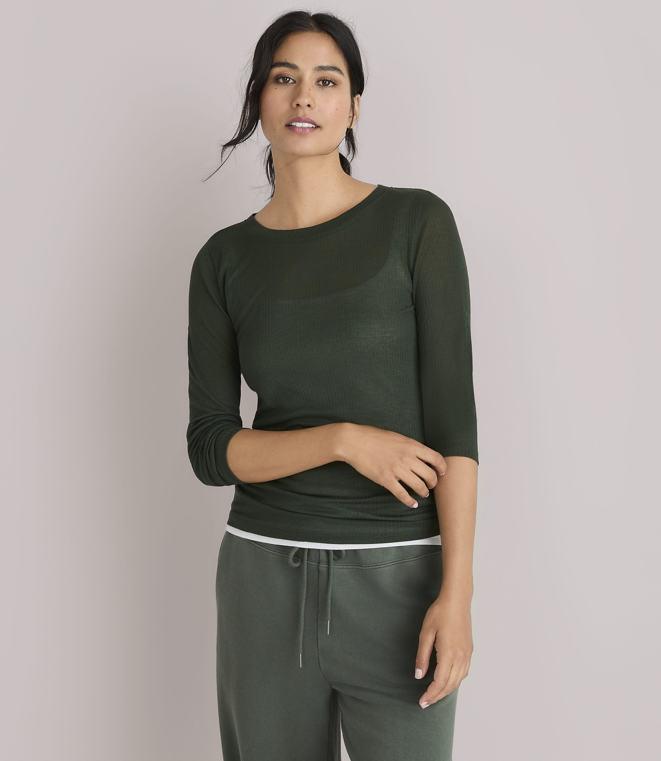 A woman in a dark green, sheer, long-sleeved shirt and green pants poses against a plain white background.