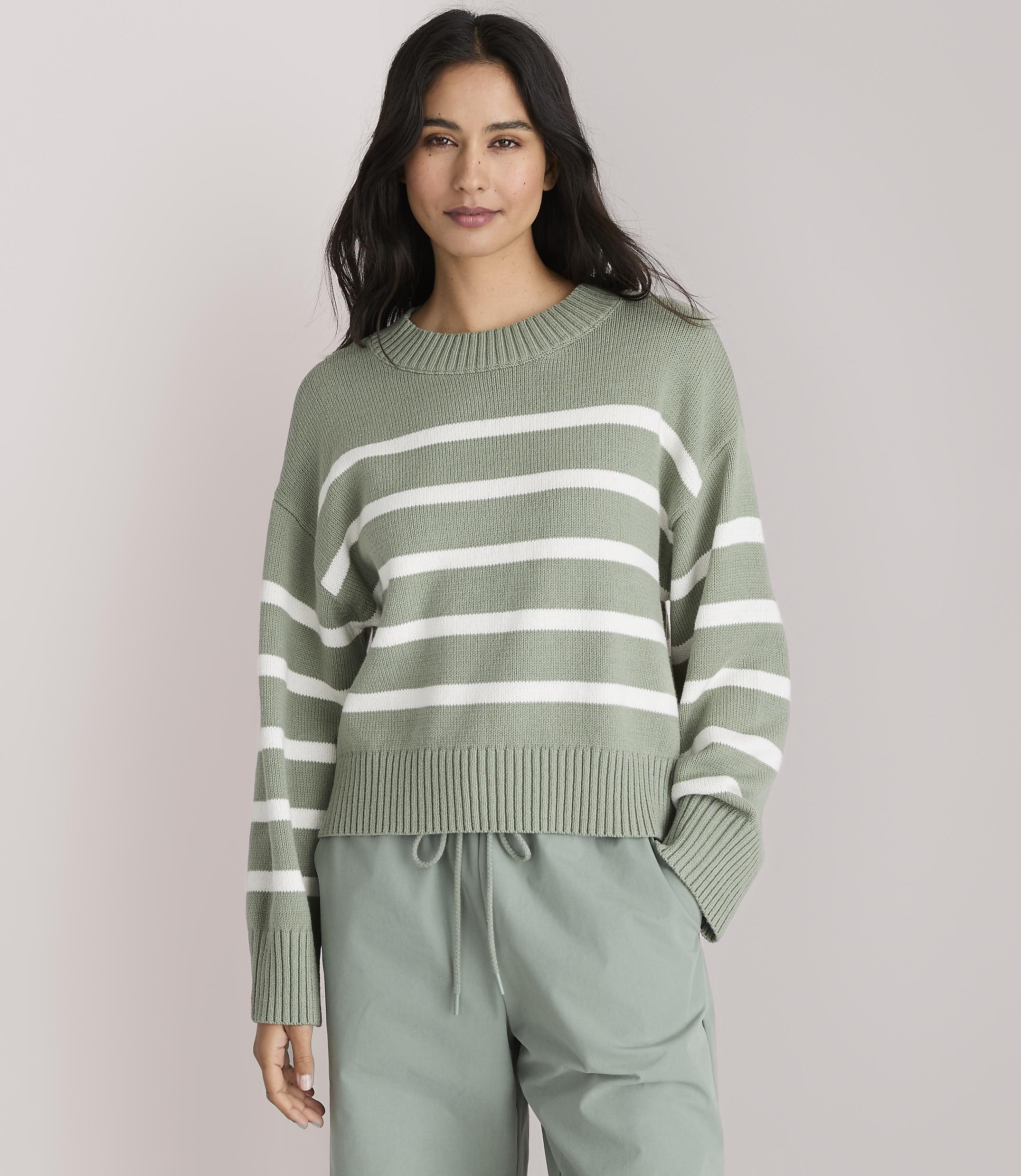 A woman in a green and white striped sweater and light green pants stands against a plain gray background.
