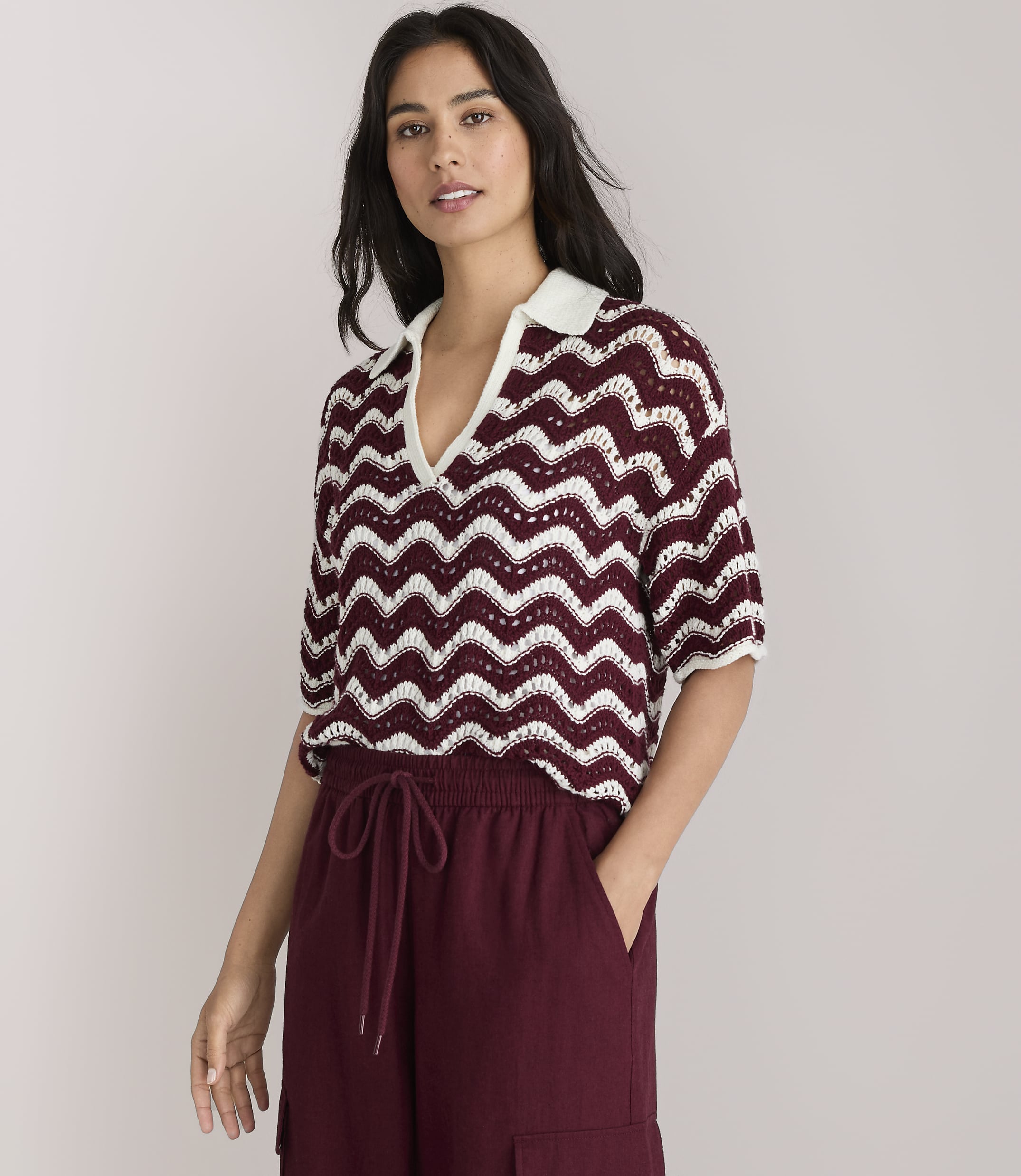 A woman in a chevron-patterned top and maroon wide-leg pants stands against a plain white background.