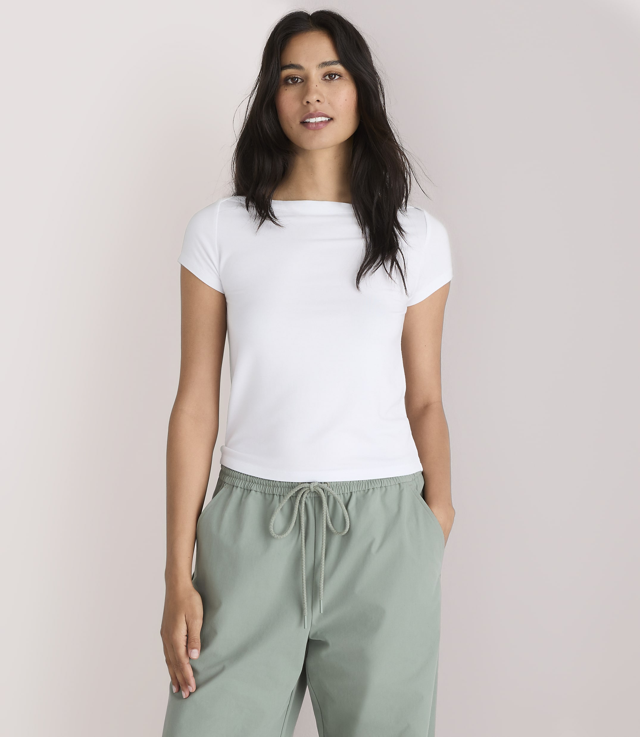 A woman in a white t-shirt and green pants stands against a plain white background.