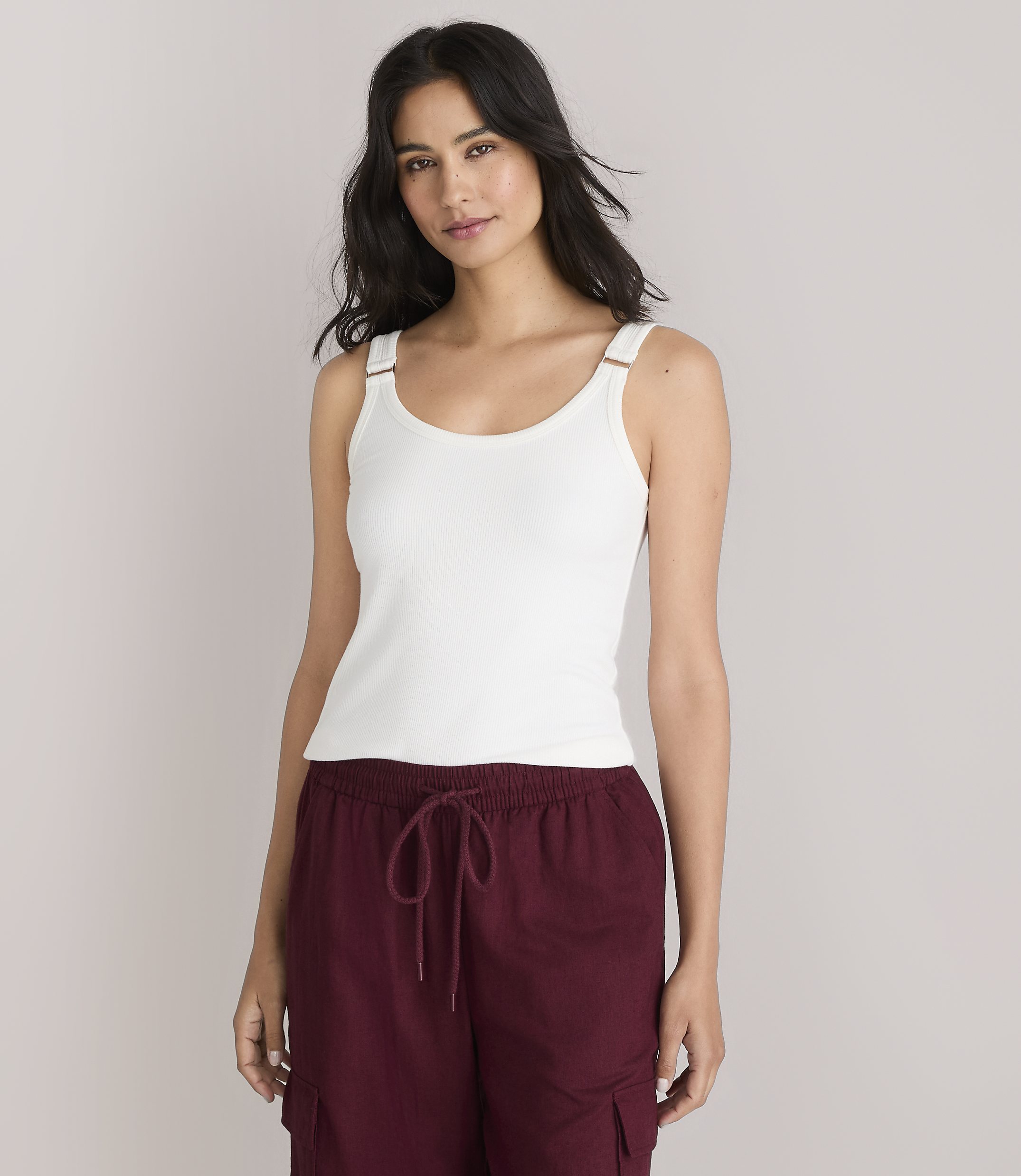A woman in a white tank top and maroon pants stands against a plain white background.
