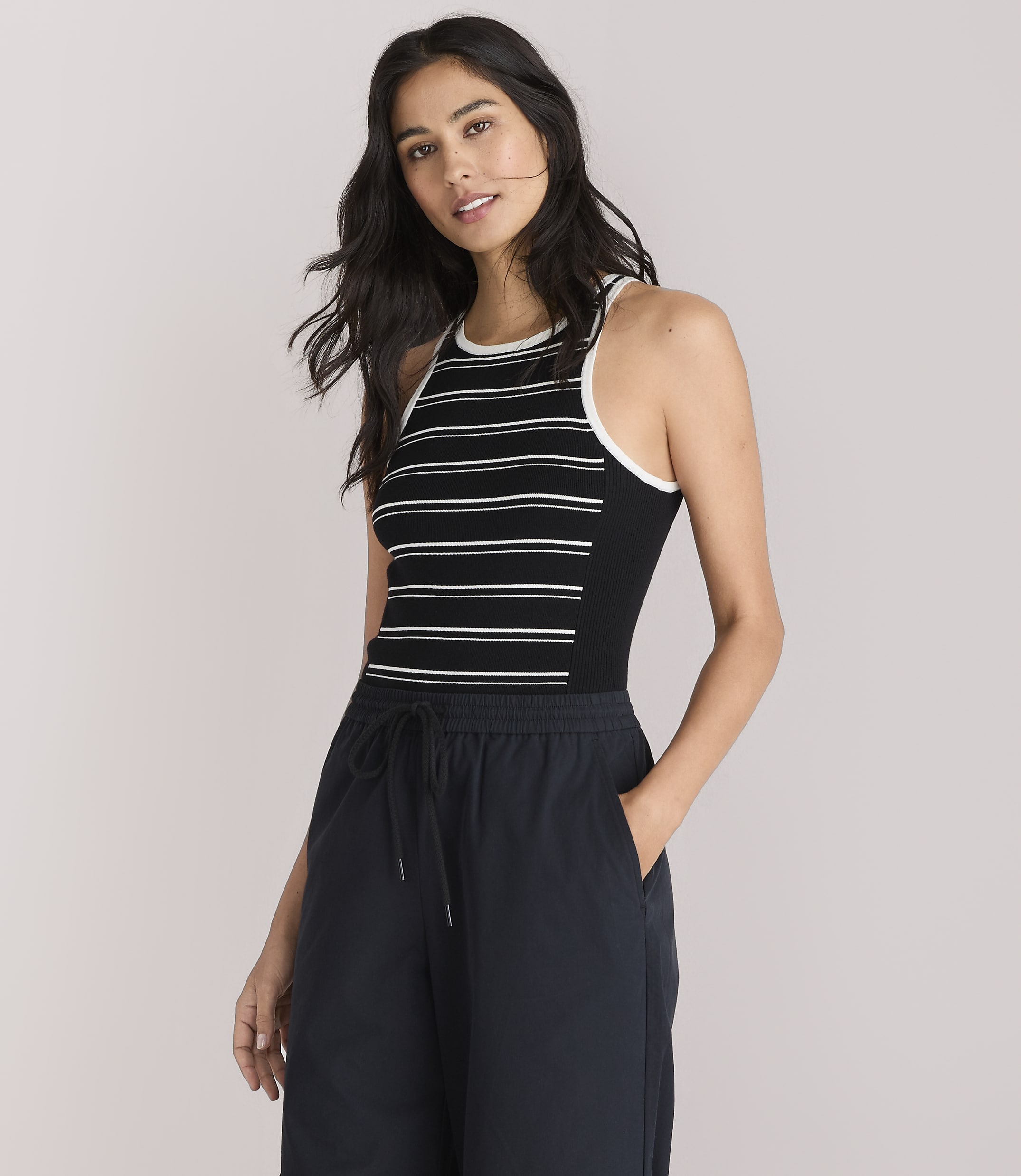 A woman in a striped tank top and dark pants stands against a plain white background.