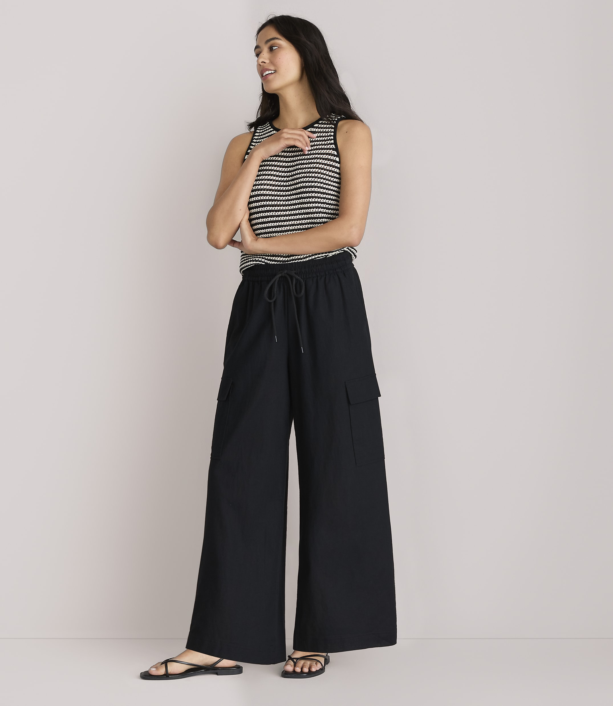 A woman in a striped tank top and black wide-leg pants stands against a plain white background.