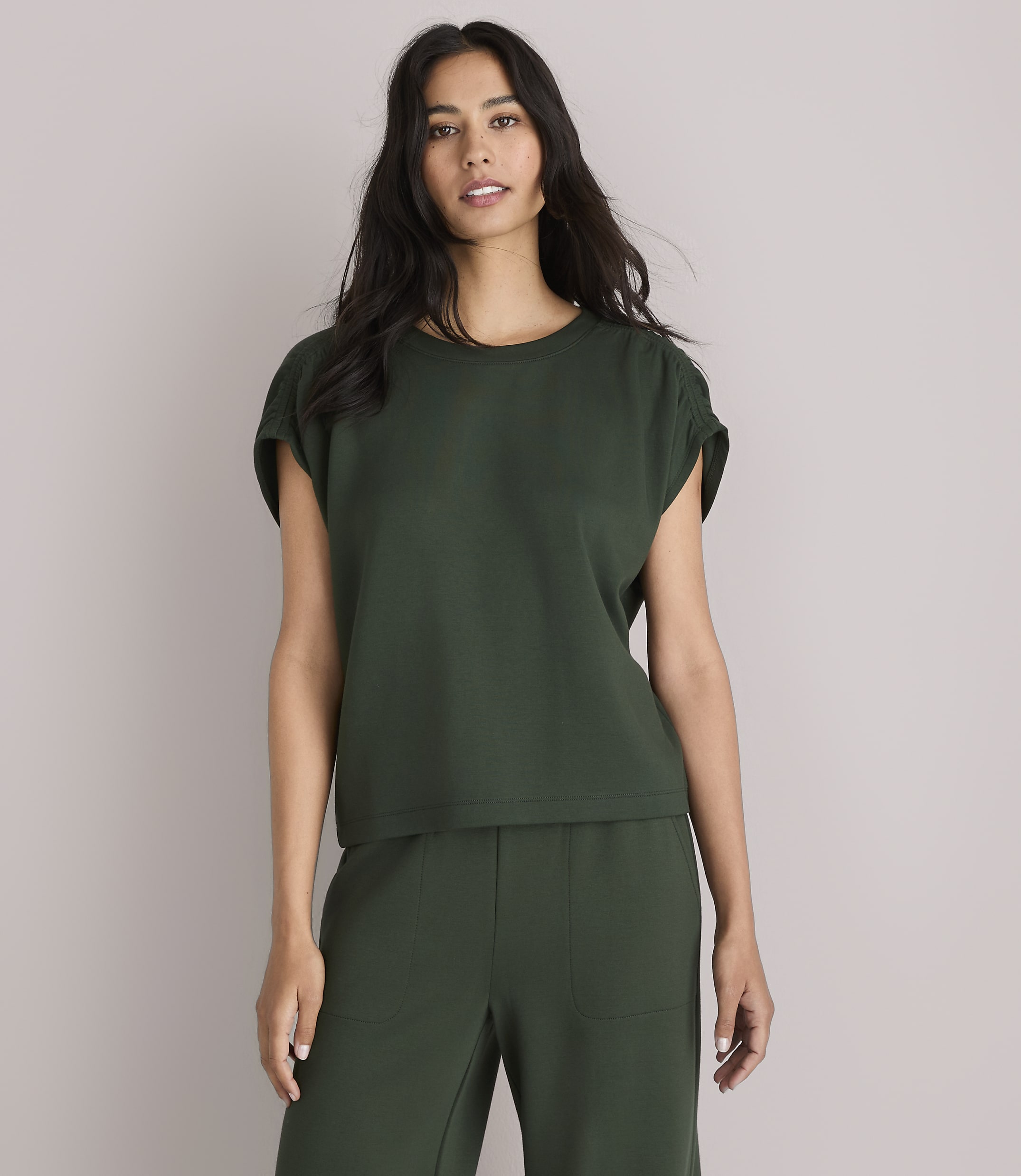 A woman in a dark green top and pants stands against a plain white background.