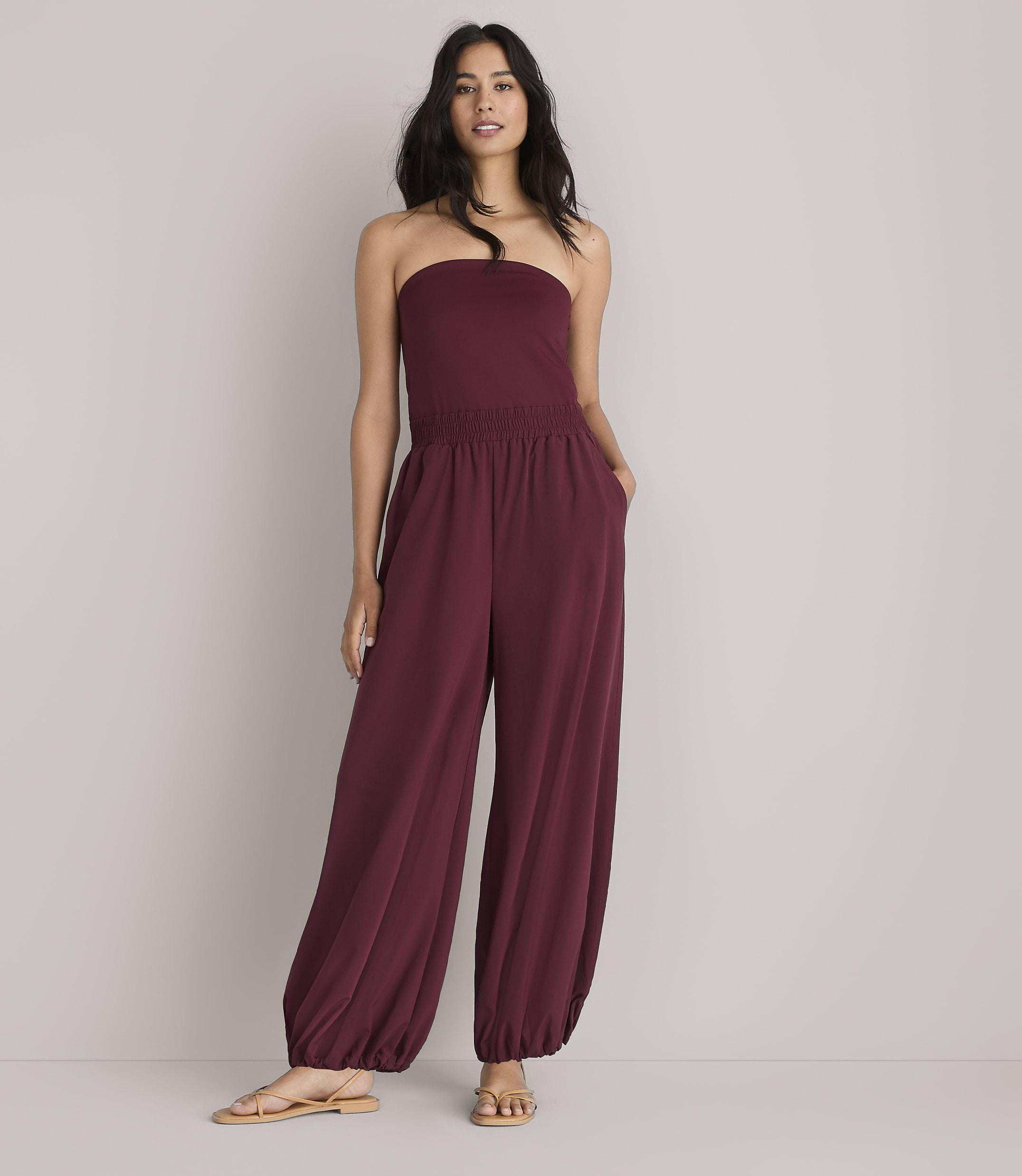 A woman in a maroon strapless jumpsuit stands against a plain white background.