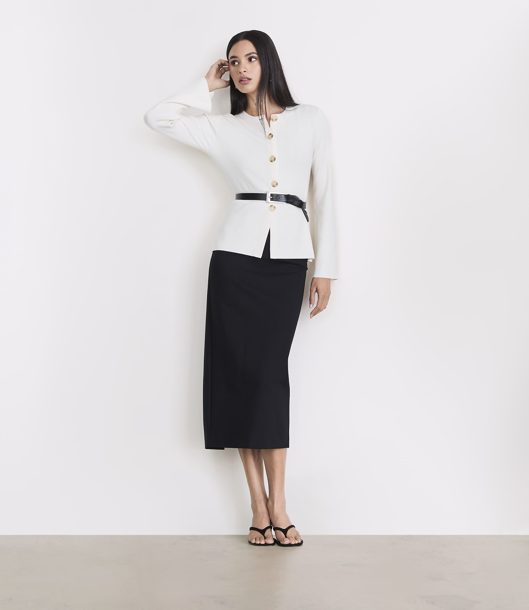 A woman in a white jacket and black skirt stands against a white wall with a light gray floor in the foreground.