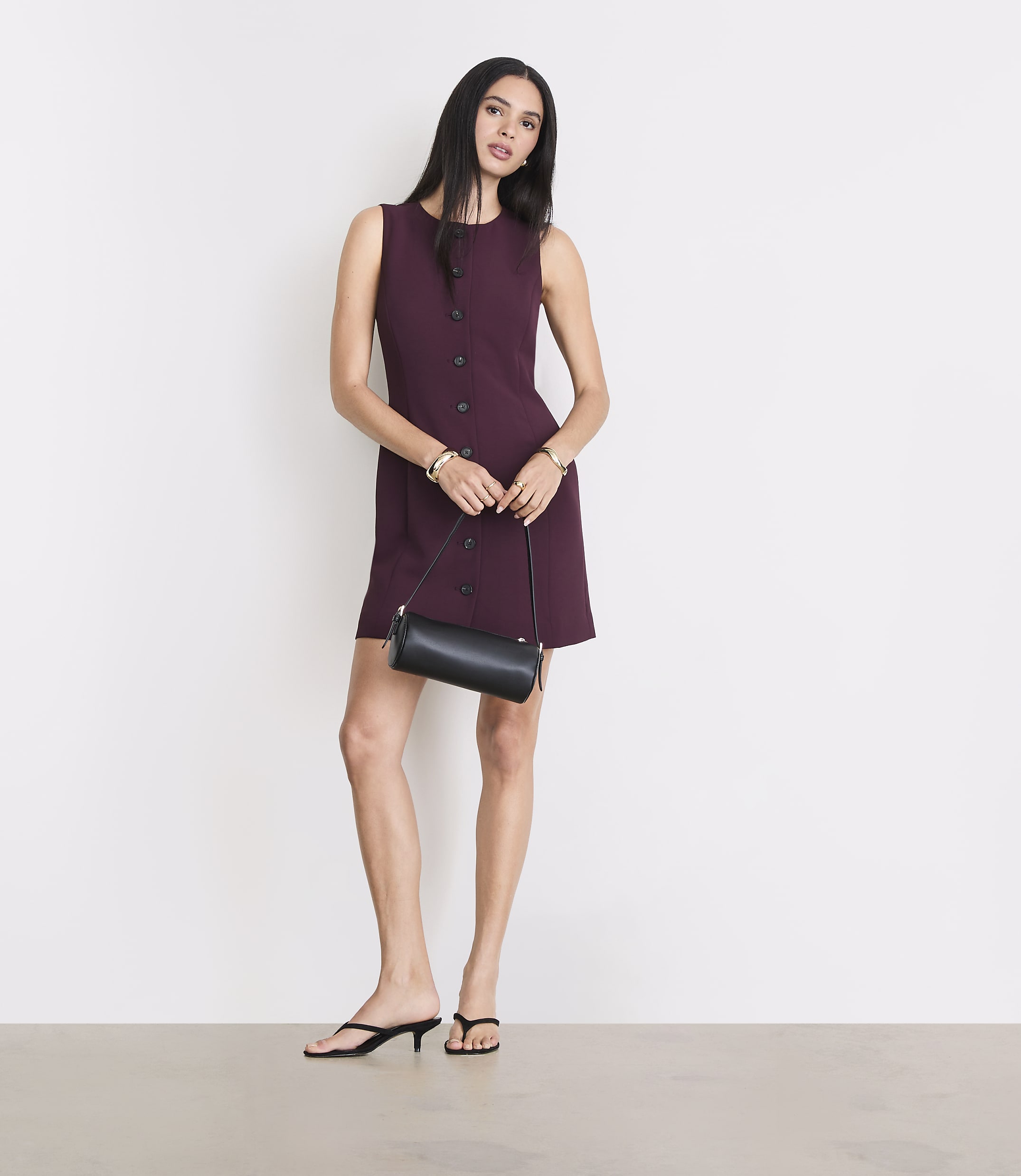 A woman in a burgundy dress and black heels stands with a black purse against a plain white background.