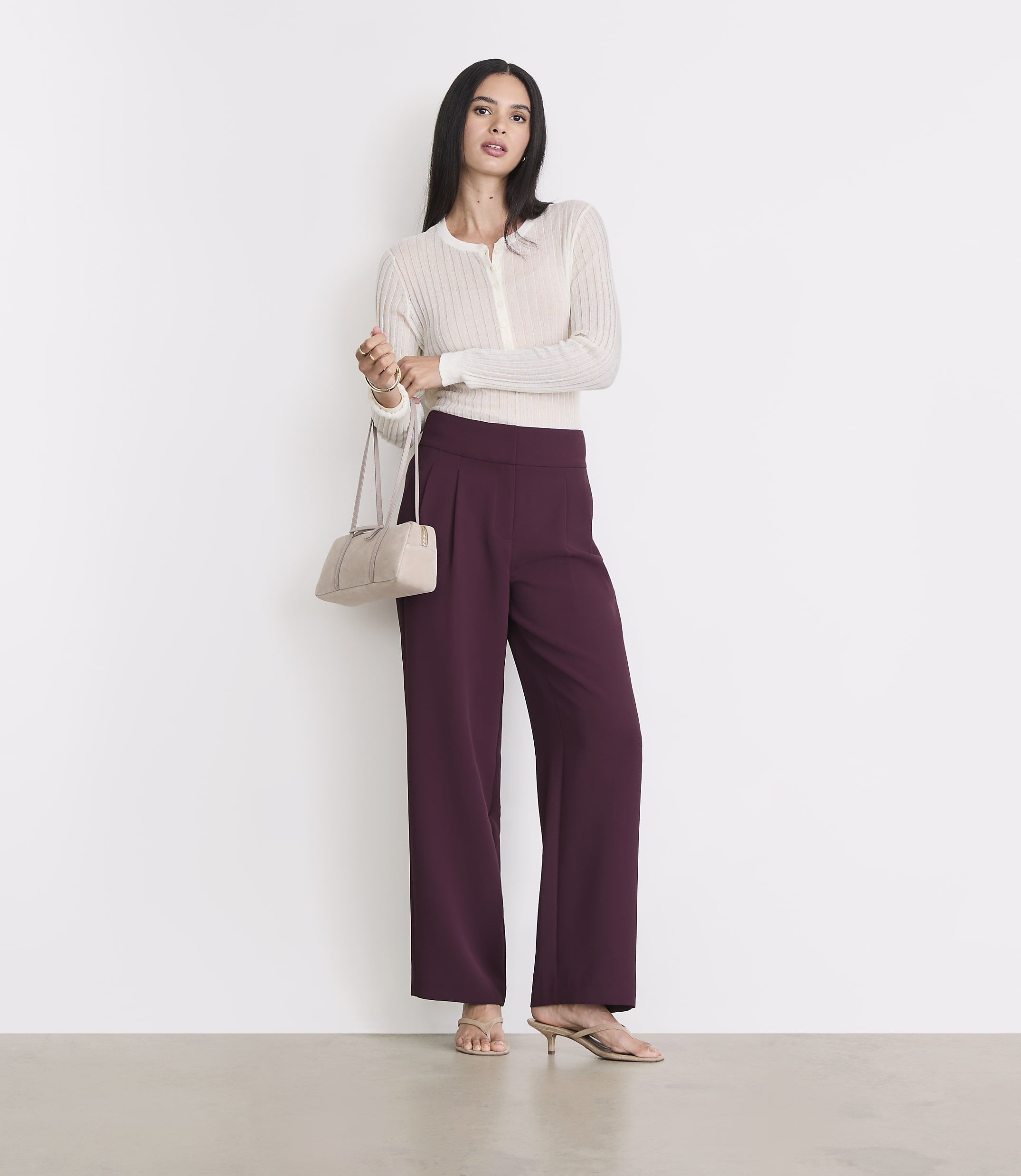 A woman in a cream cardigan and plum wide-leg pants stands against a white background, holding a cream handbag.