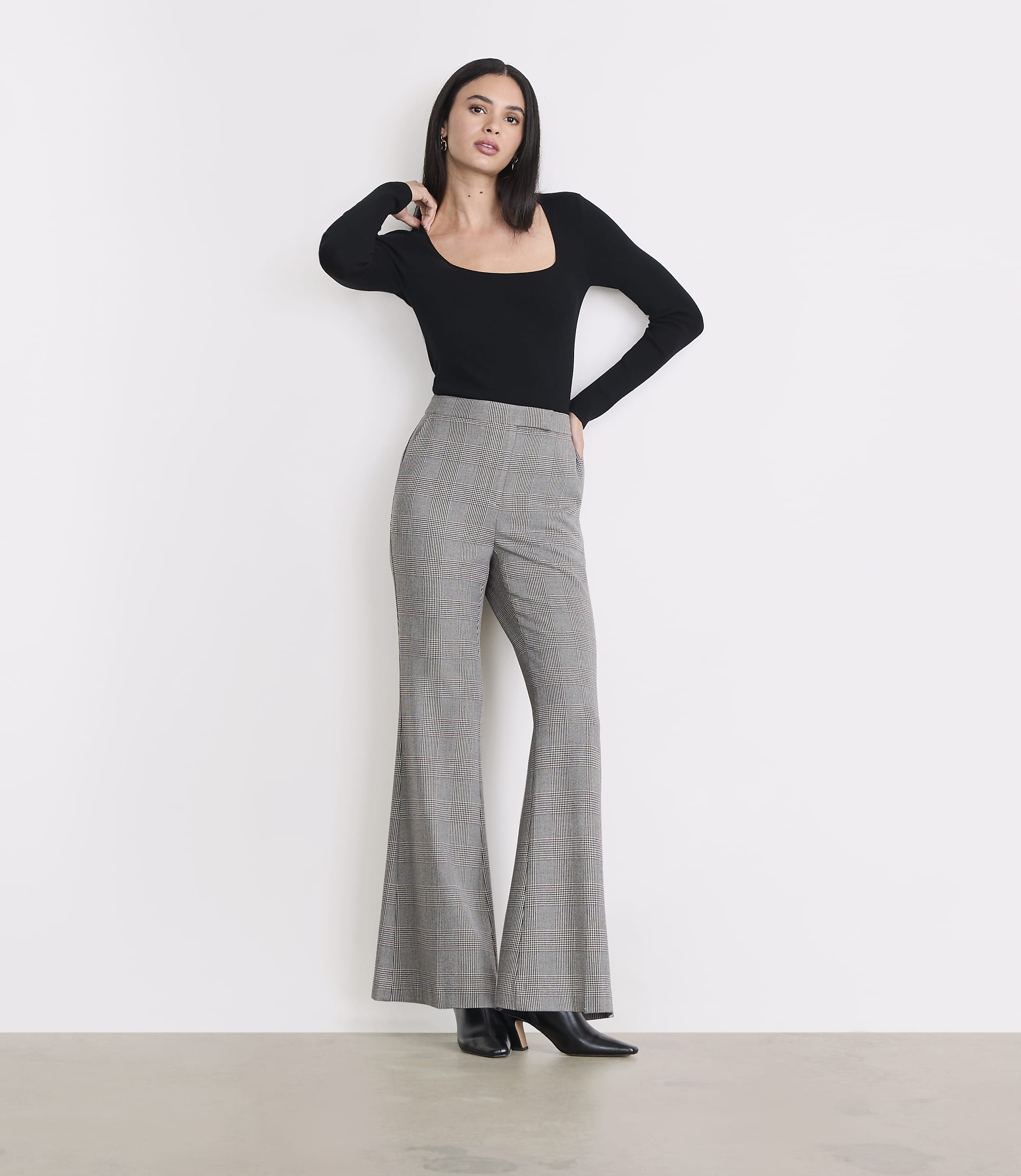 A woman in a black top and plaid bell-bottom pants stands against a plain white background.