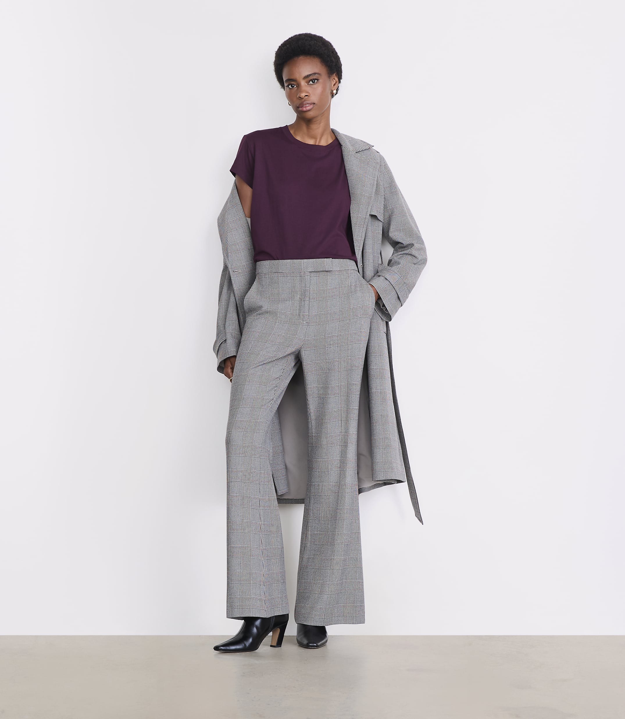A woman in a plum t-shirt, plaid wide-leg pants, and a grey trench coat stands against a plain white background.