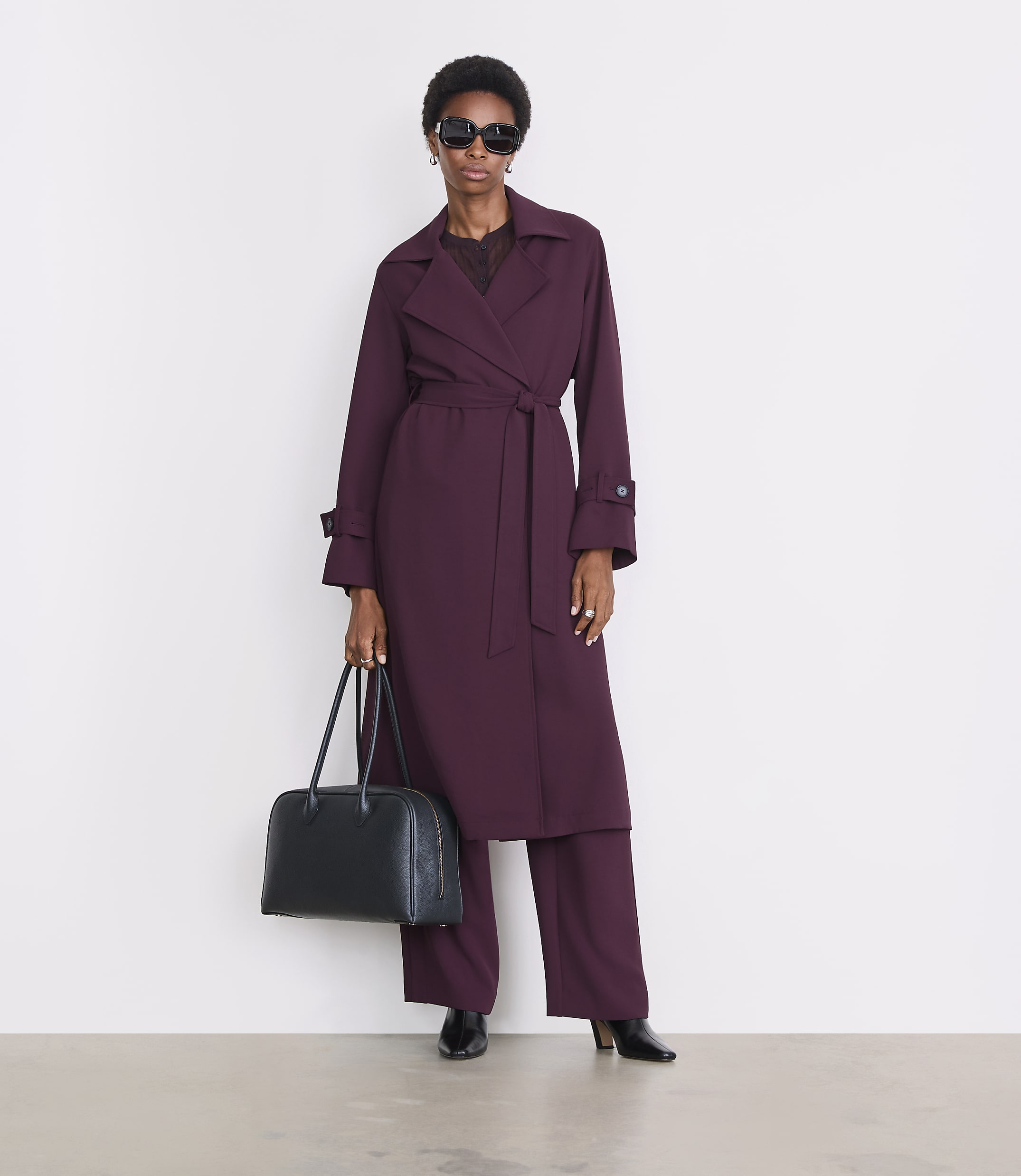 A woman in a plum trench coat and matching pants stands with a dark handbag against a plain white background.