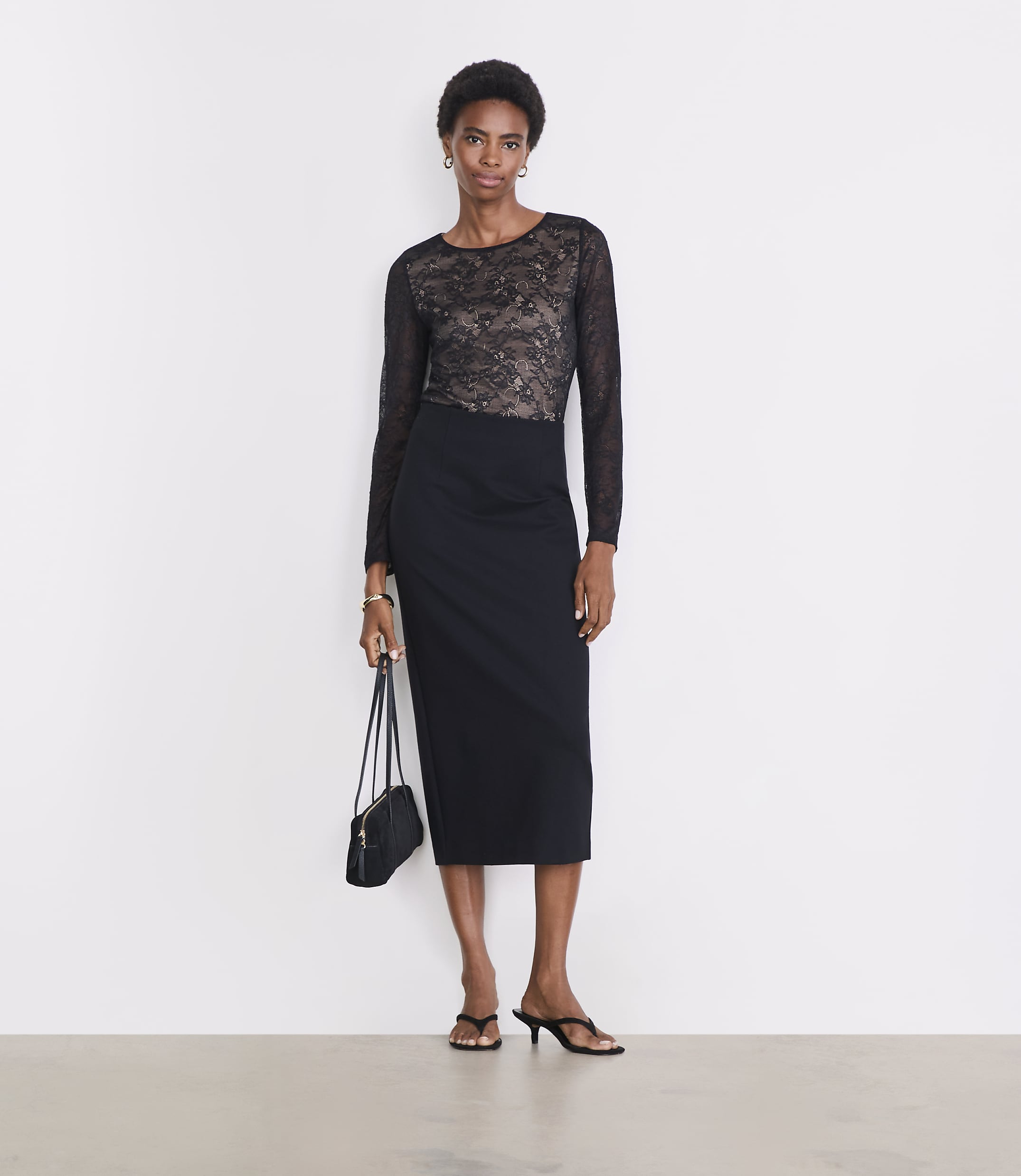 A woman in a black lace top and pencil skirt stands against a plain white background, holding a small black purse.
