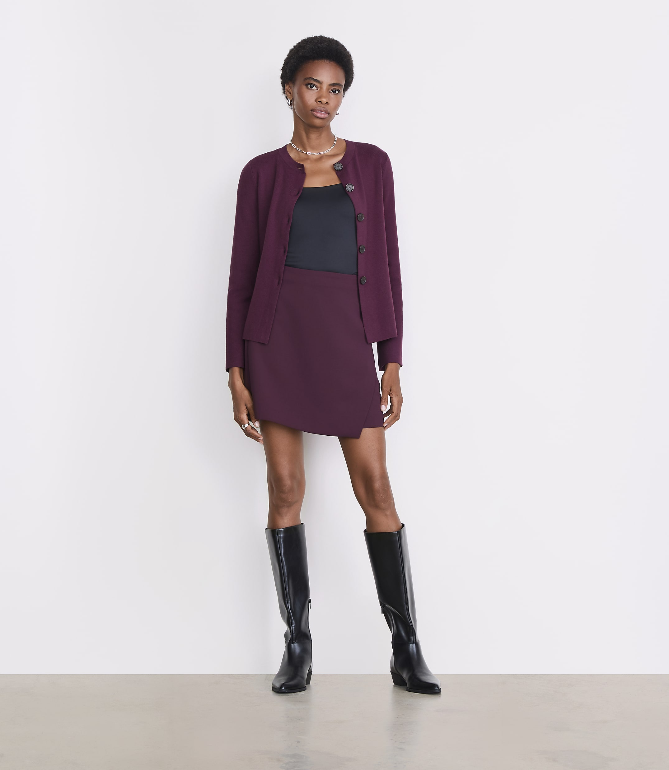 A woman in a burgundy cardigan, black top, and matching skirt stands in front of a plain white background.