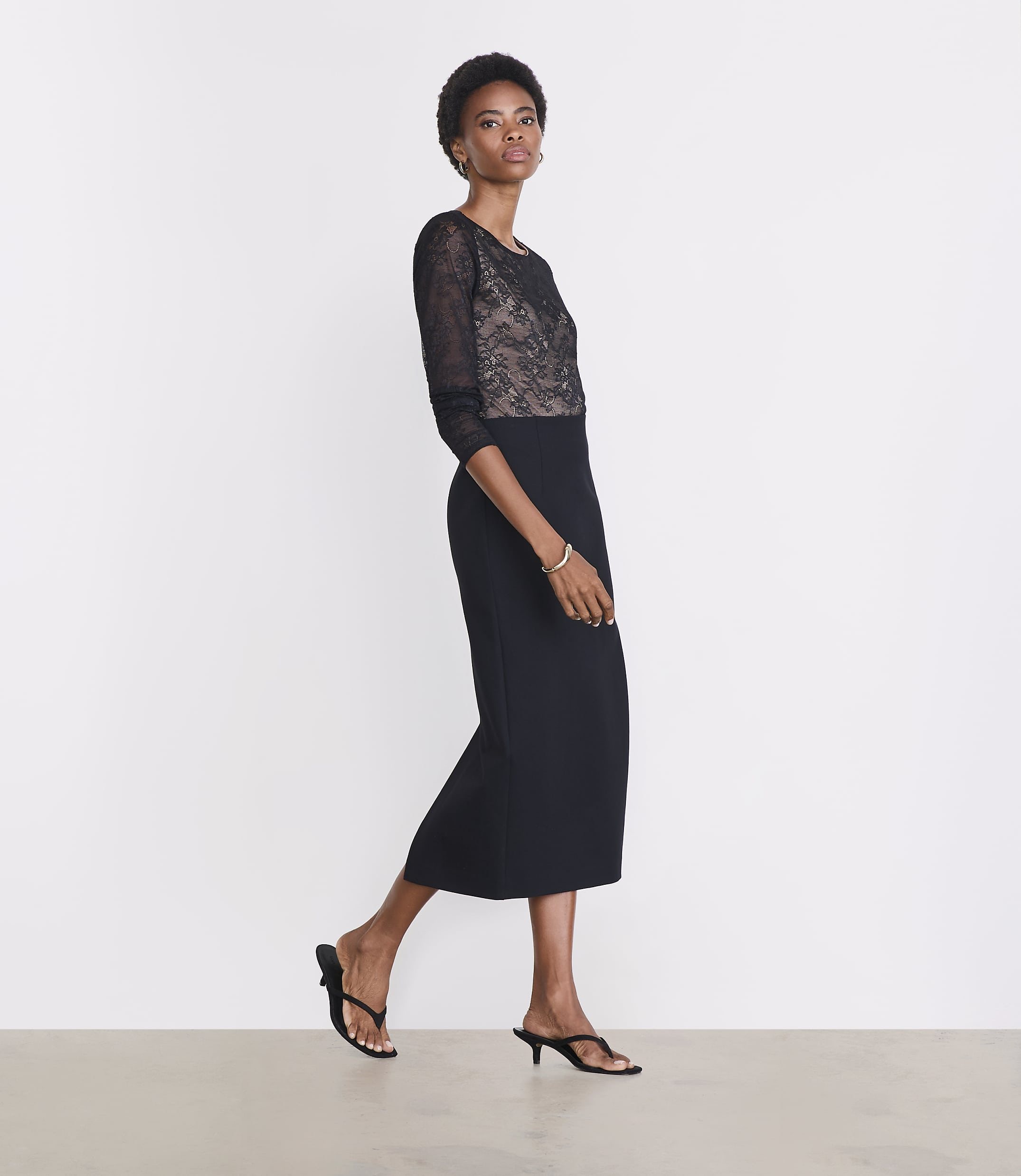 A woman in a black lace-bodice dress and heeled sandals stands against a plain white background.