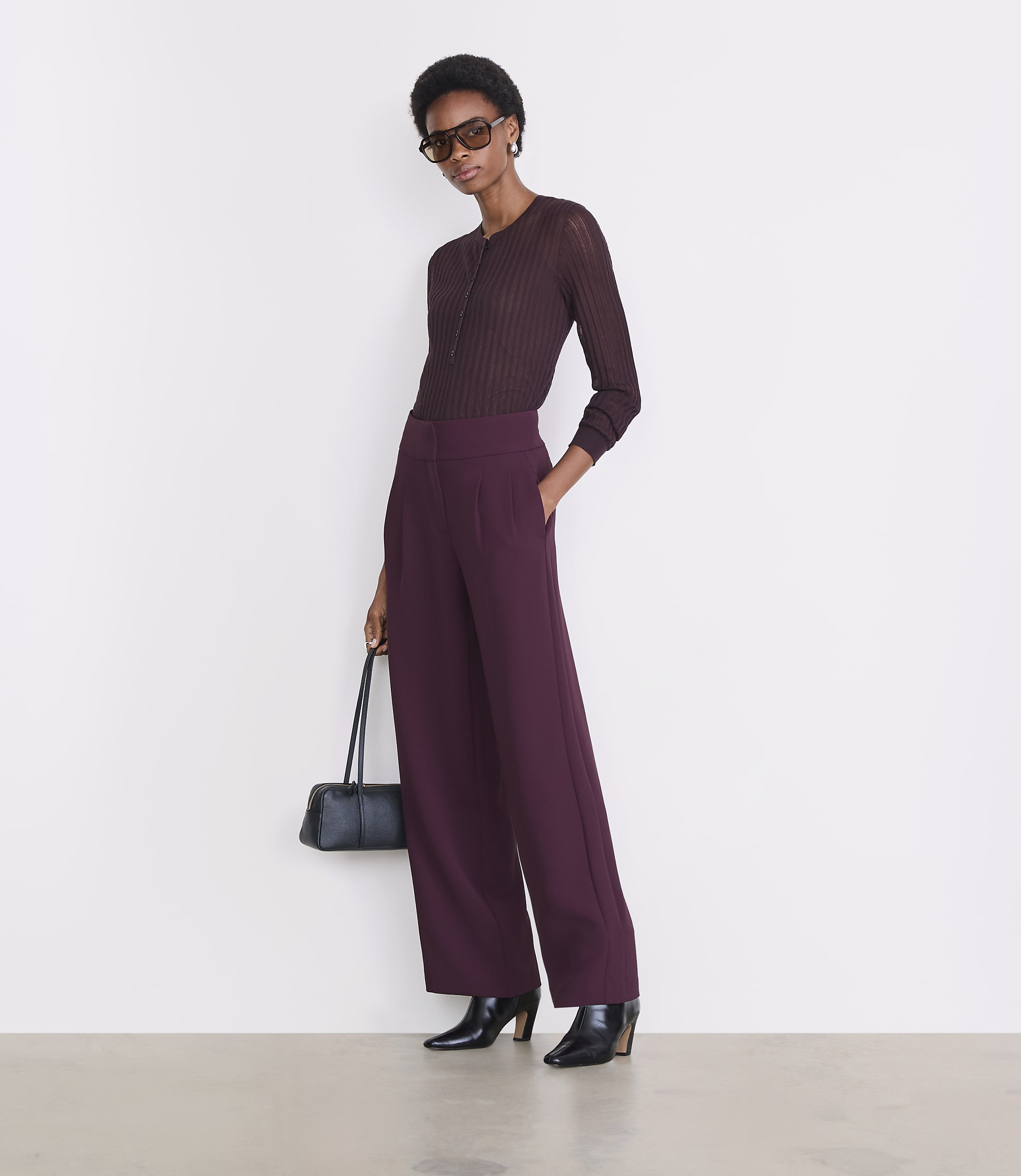 A woman in a maroon top and wide-leg maroon pants stands against a plain white background, holding a black handbag.