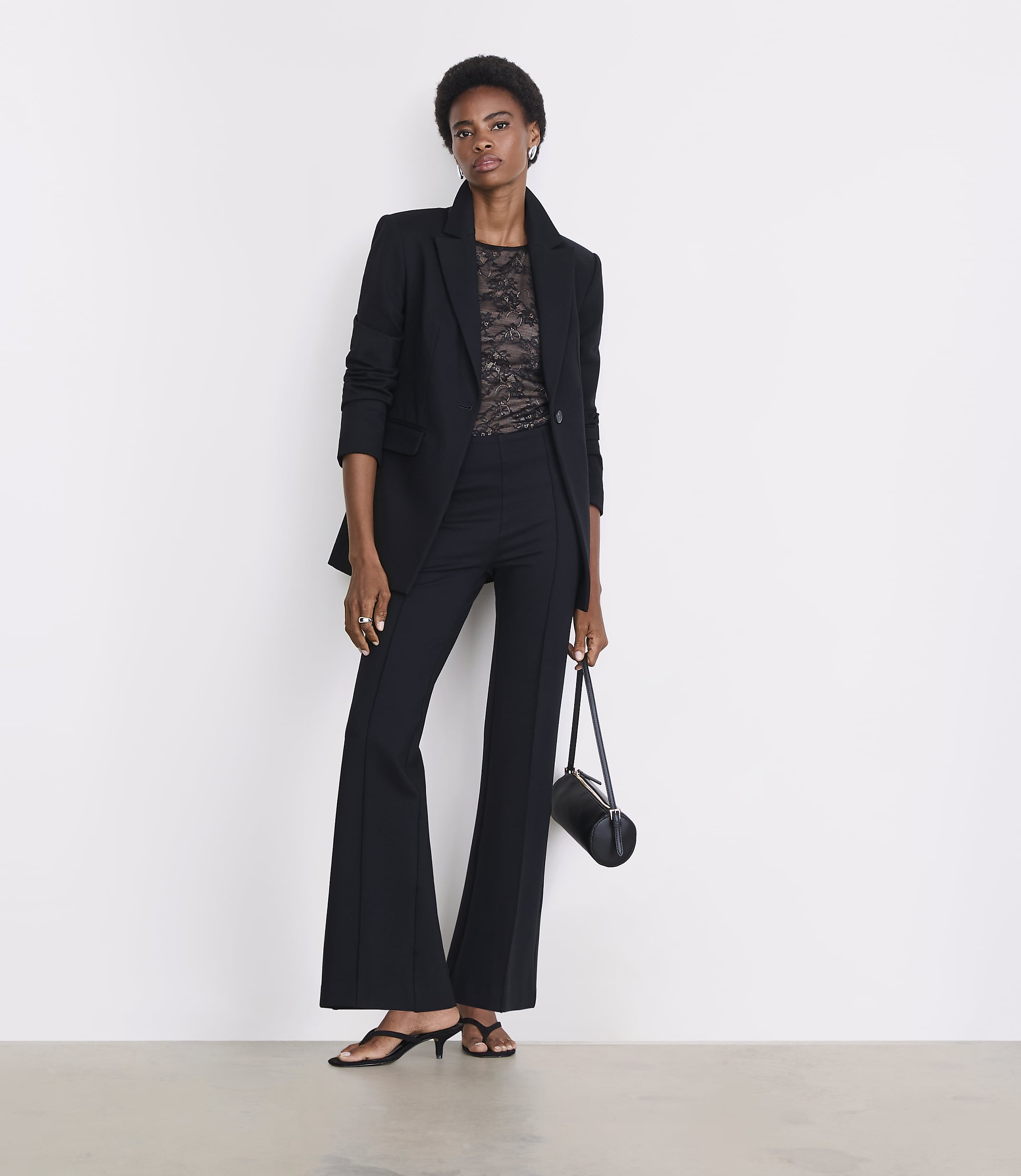 A woman in a black blazer and flared pants stands against a plain white background, holding a small black bag.