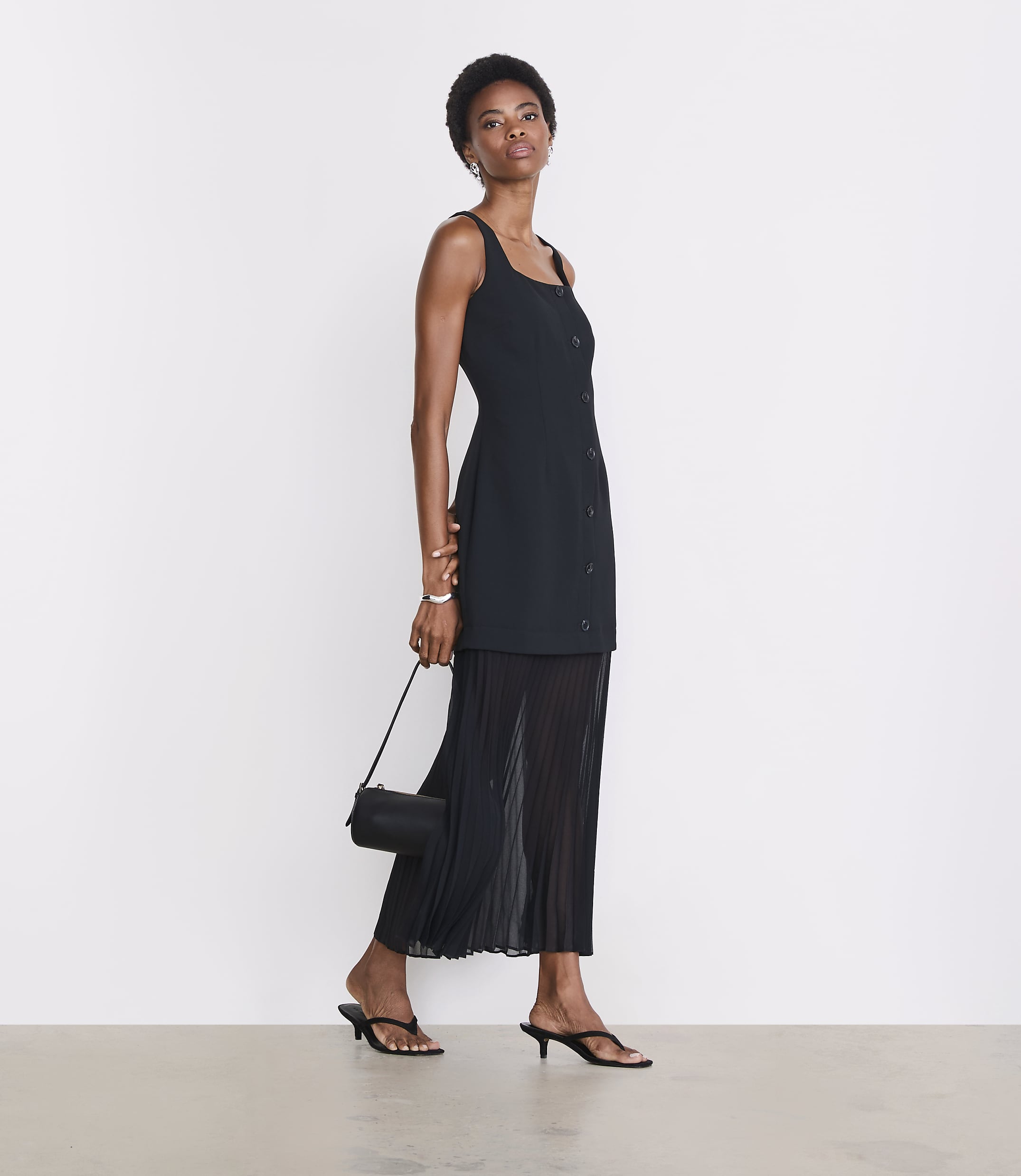 A woman in a black dress and sandals stands against a plain white background, holding a small black purse.