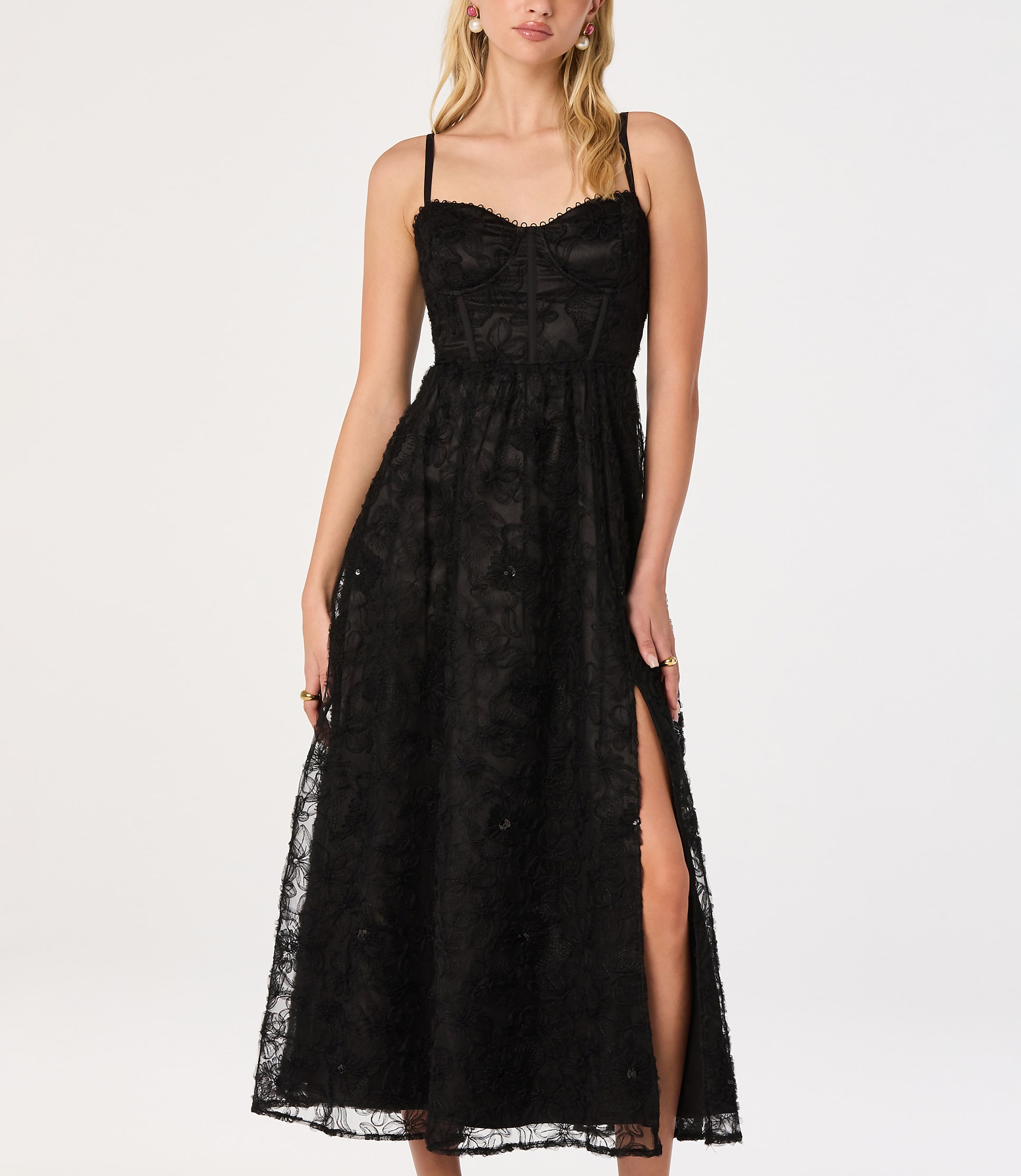 A woman in a black lace dress with a thigh-high slit stands against a plain white background.