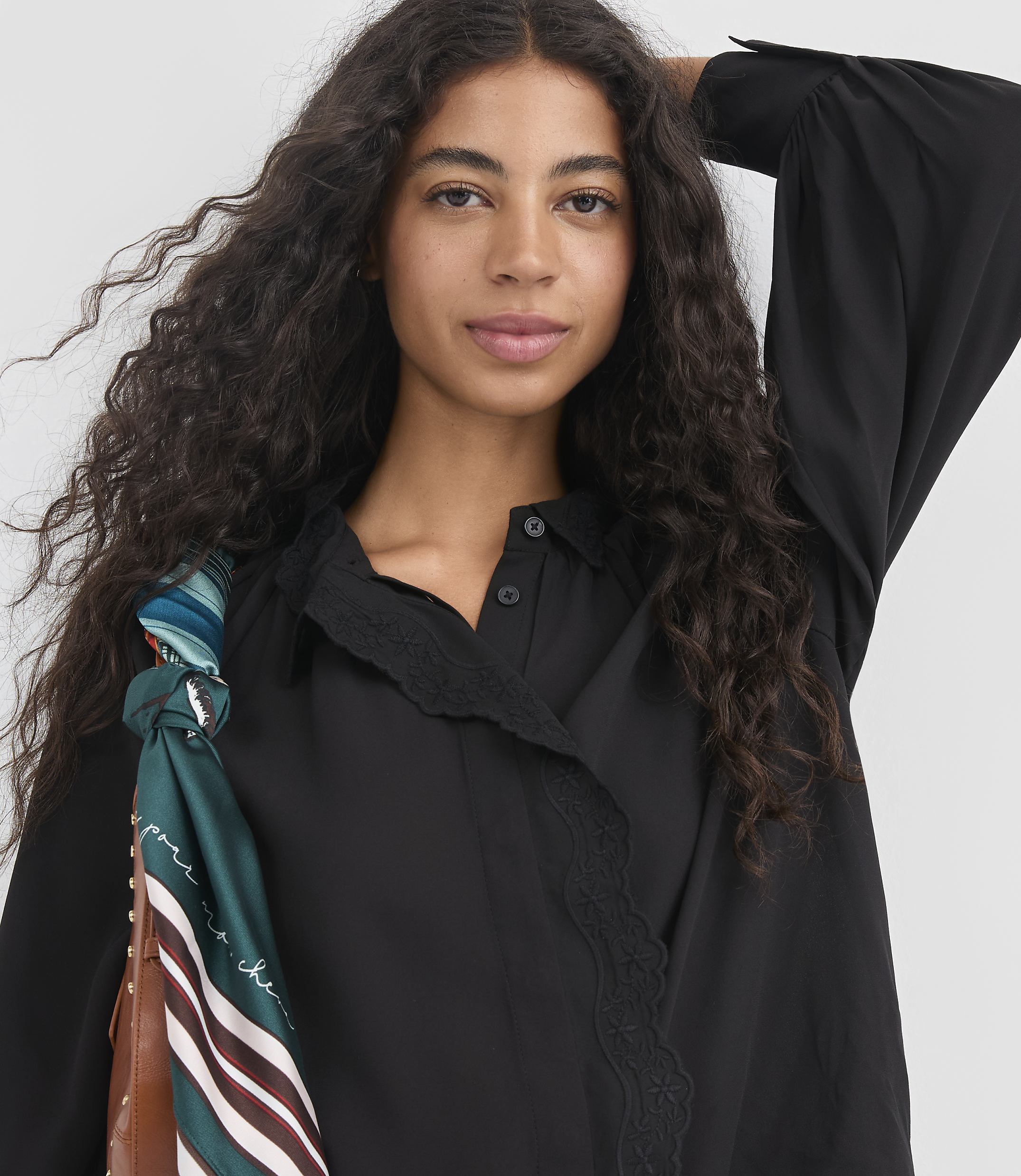 A woman with long, dark, curly hair wears a black blouse with lace trim, and a patterned scarf is tied to her brown bag in the foreground, against a plain background.