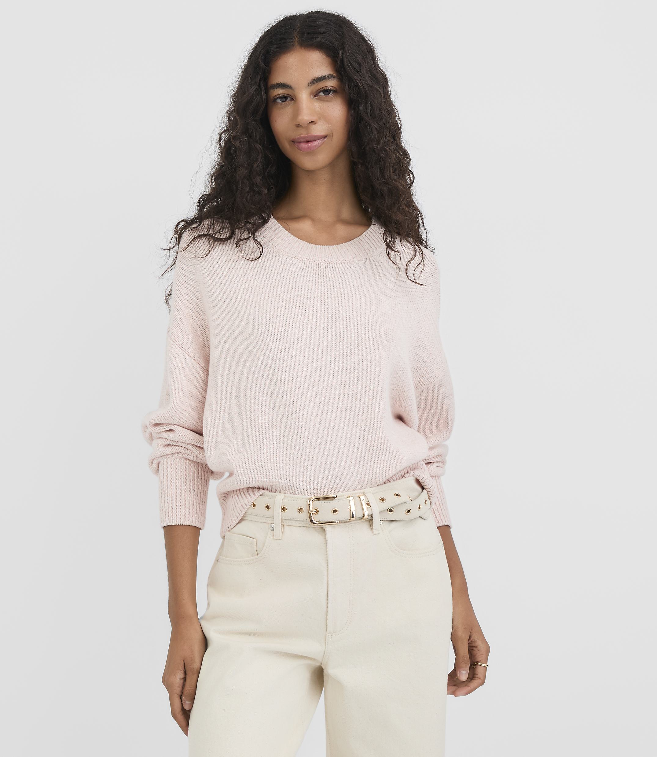 A woman in a light pink sweater and cream pants stands against a plain white background.