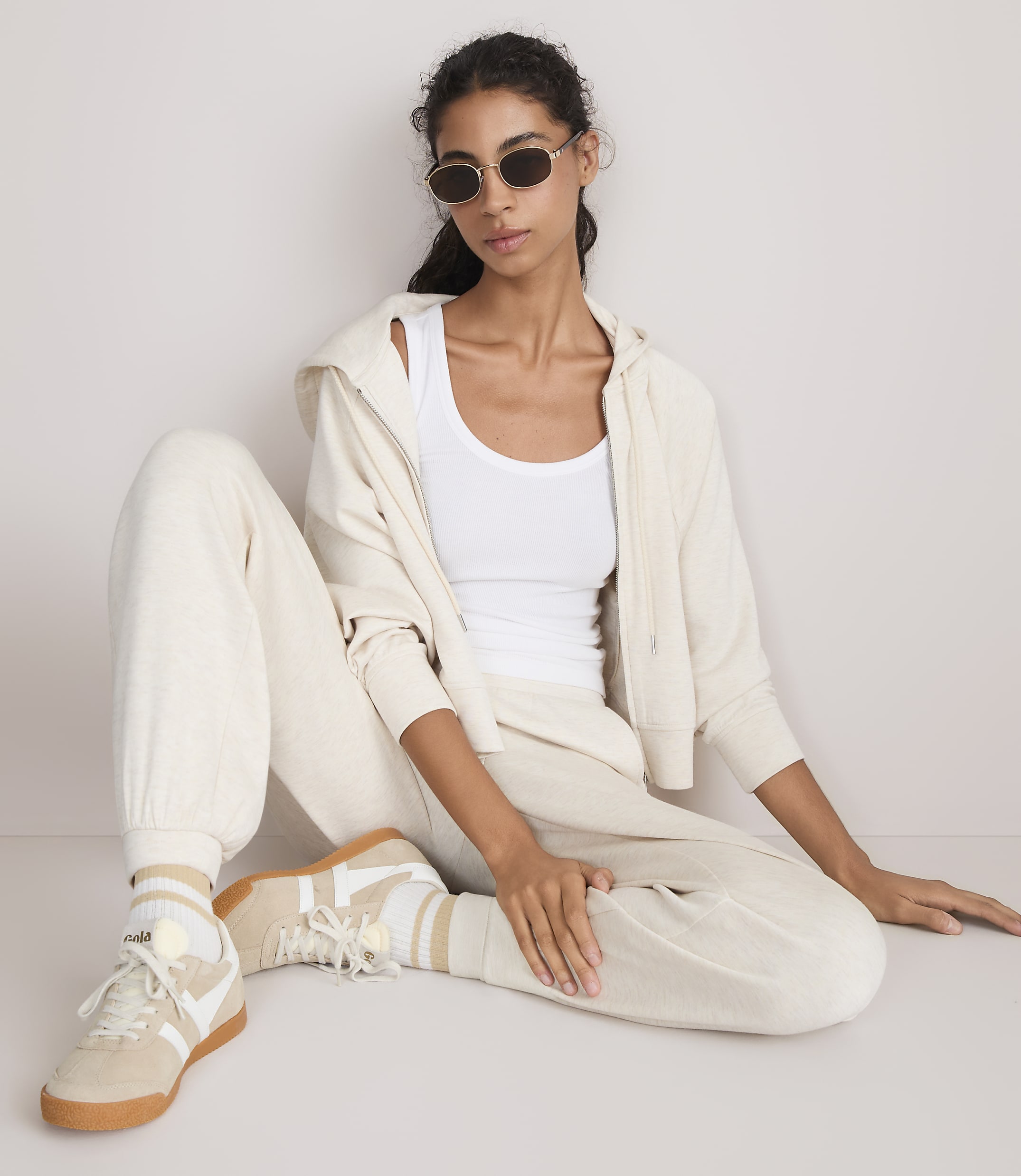 A woman in a white tank top, cream hoodie, and matching sweatpants sits on a light-colored floor, her legs crossed and wearing sneakers, against a plain, light-colored background.