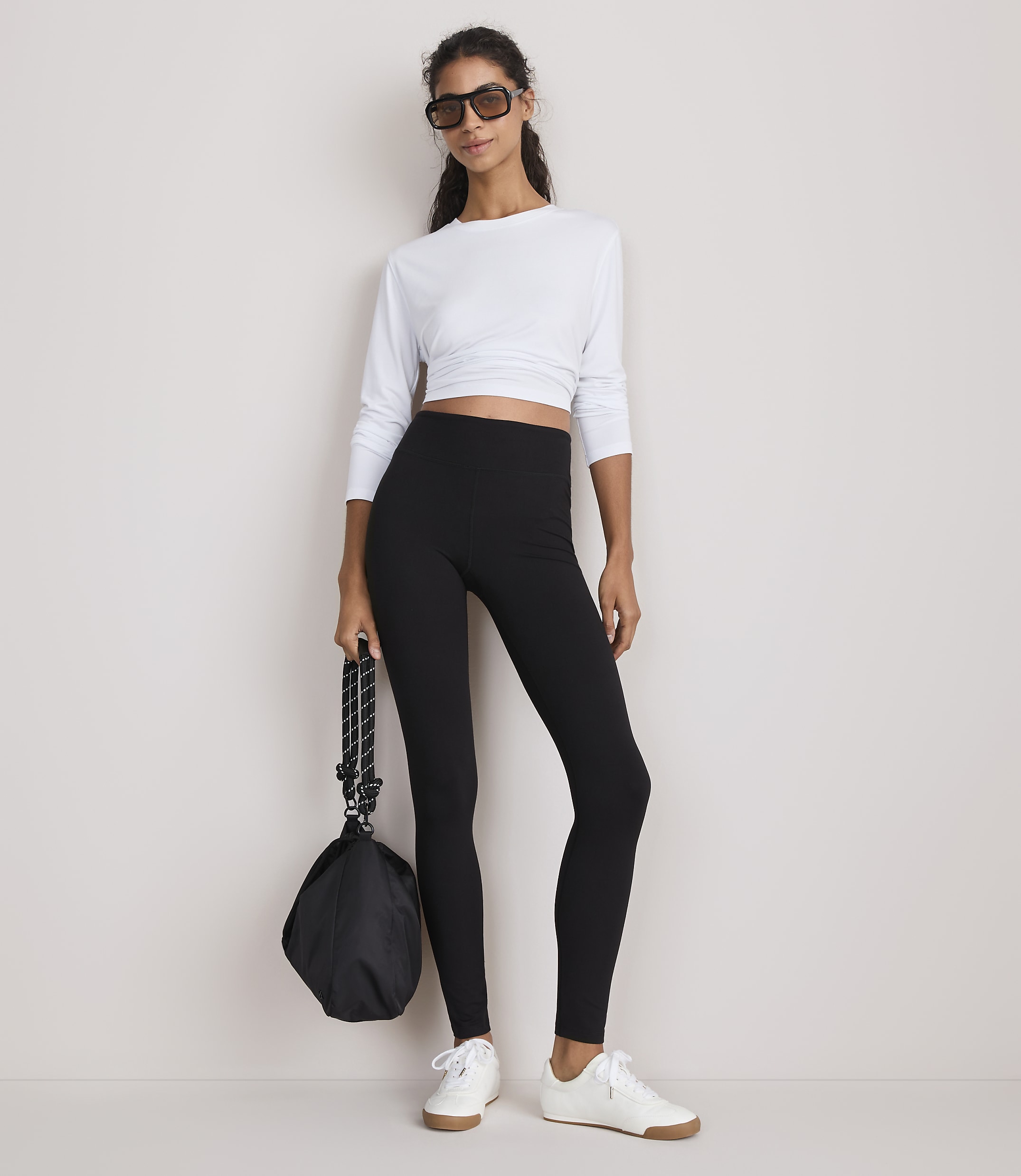 A woman in a white long-sleeved crop top and black leggings stands against a plain white background, holding a black bag.