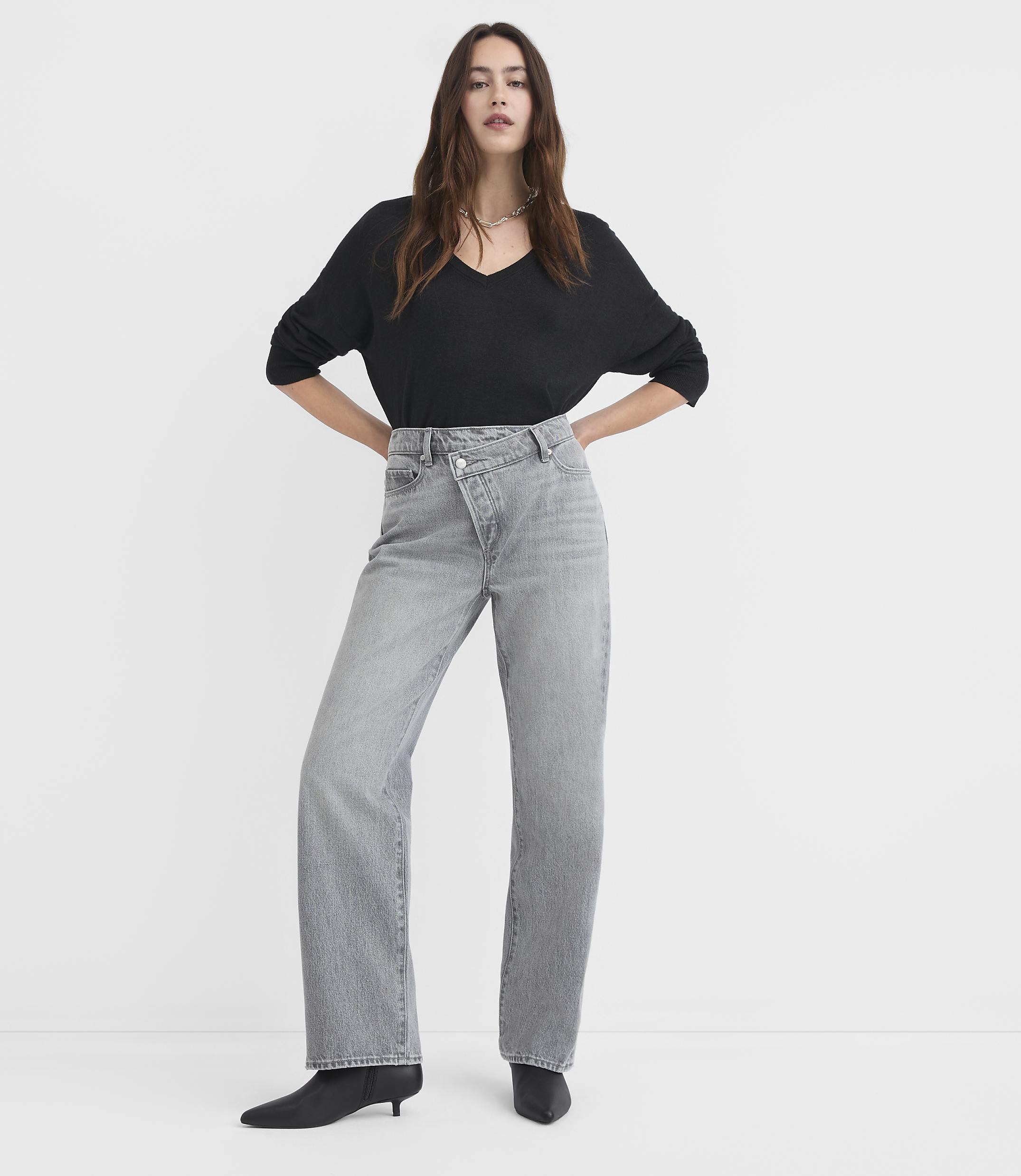 A woman in a black v-neck shirt and grey criss-cross jeans stands against a plain white background.