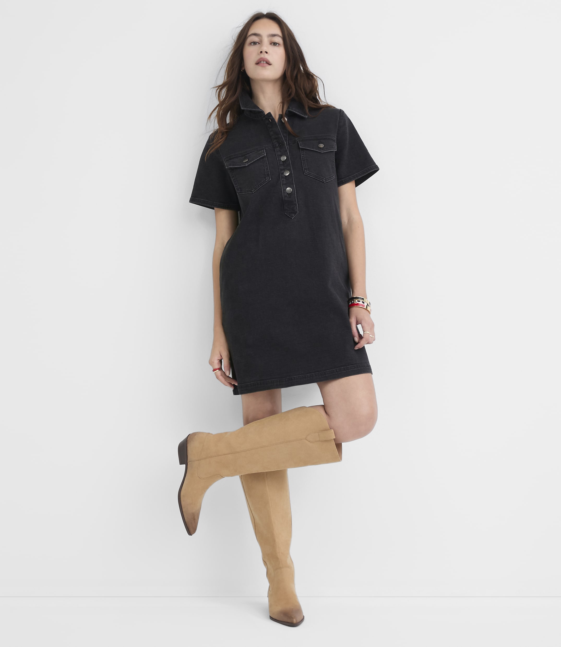 A woman in a black denim dress and tan boots stands against a plain white background.