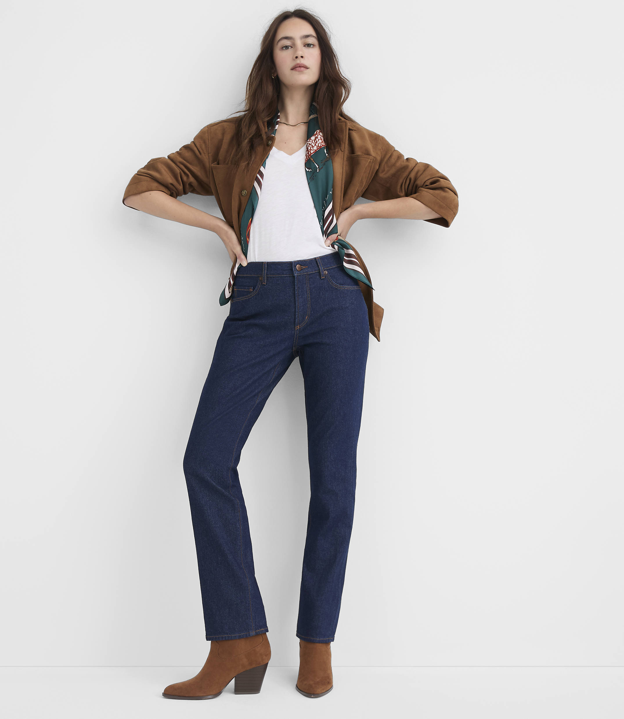 A woman in a brown jacket, white top, and dark jeans stands against a plain white background.