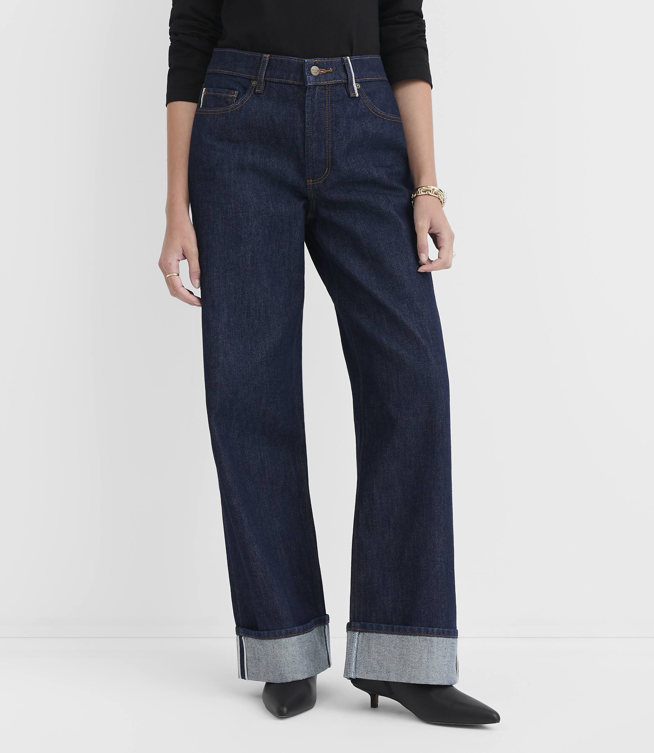 A person stands in dark wash, wide-leg jeans with cuffed hems and black heels against a plain white background.