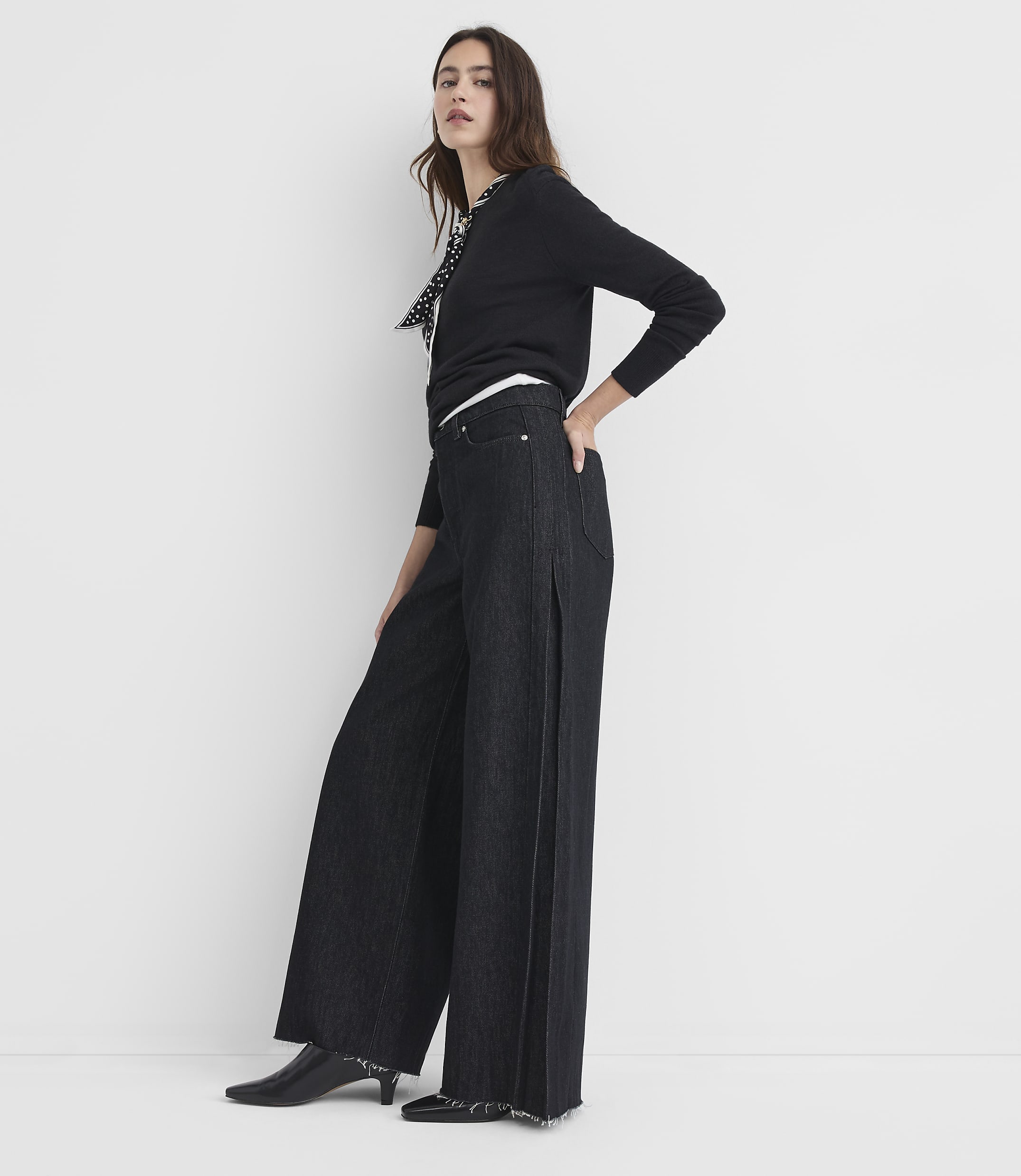 A woman in a black sweater and wide-leg jeans stands against a plain white background.
