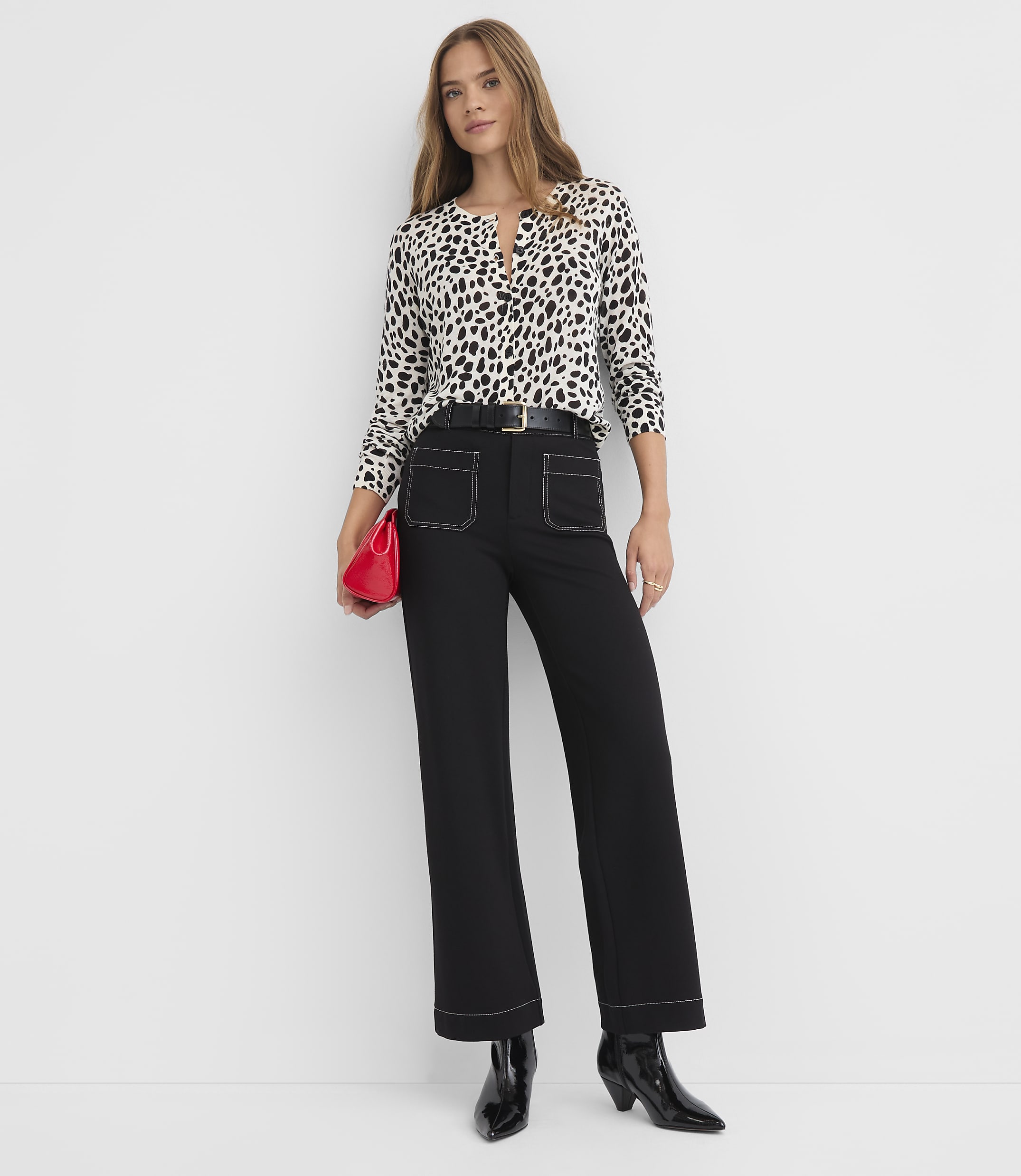 A woman in a black and white spotted blouse and black wide-leg pants stands against a plain white background, holding a red clutch.