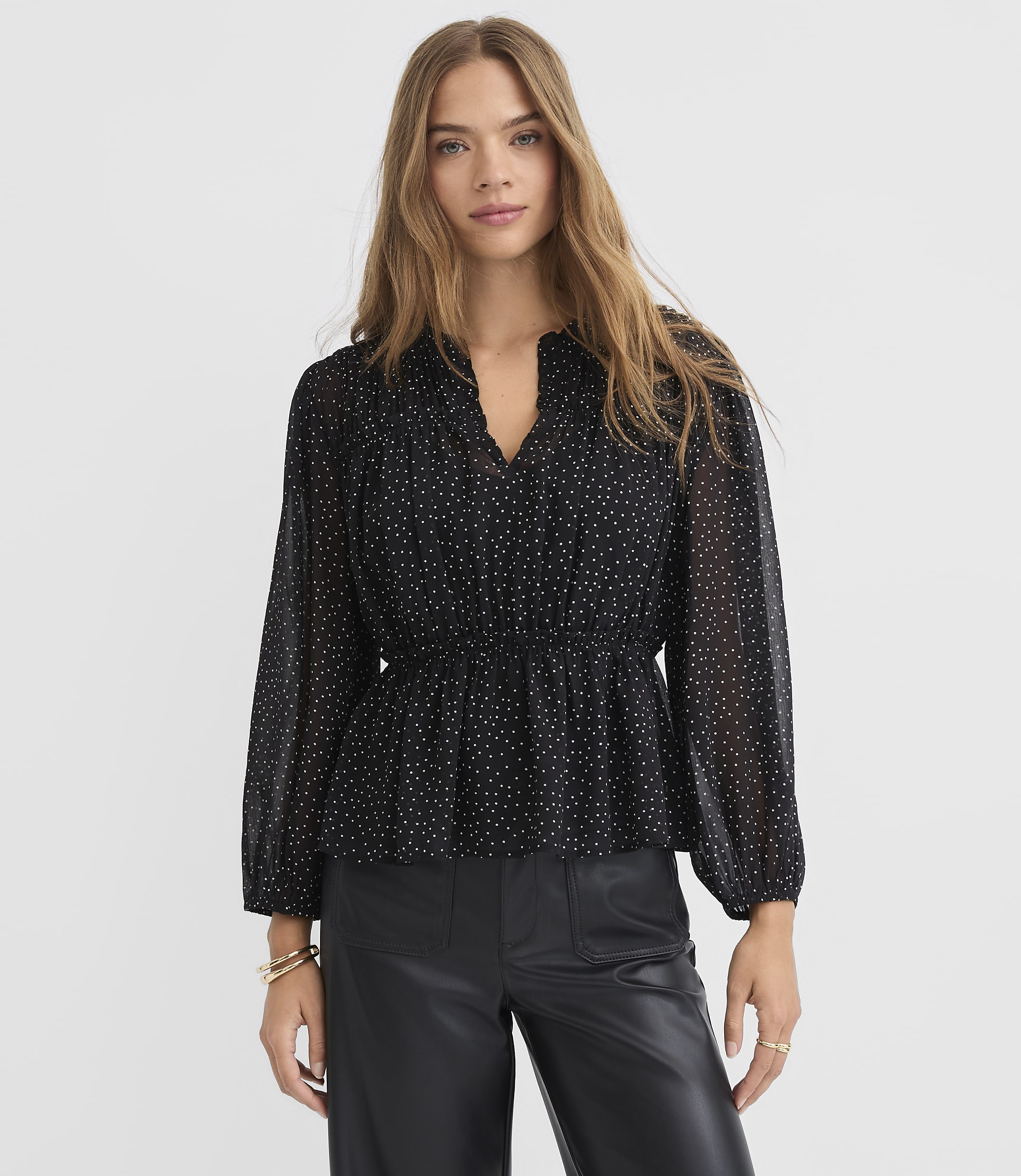 A woman in a black polka dot blouse and leather pants stands against a white textured background.