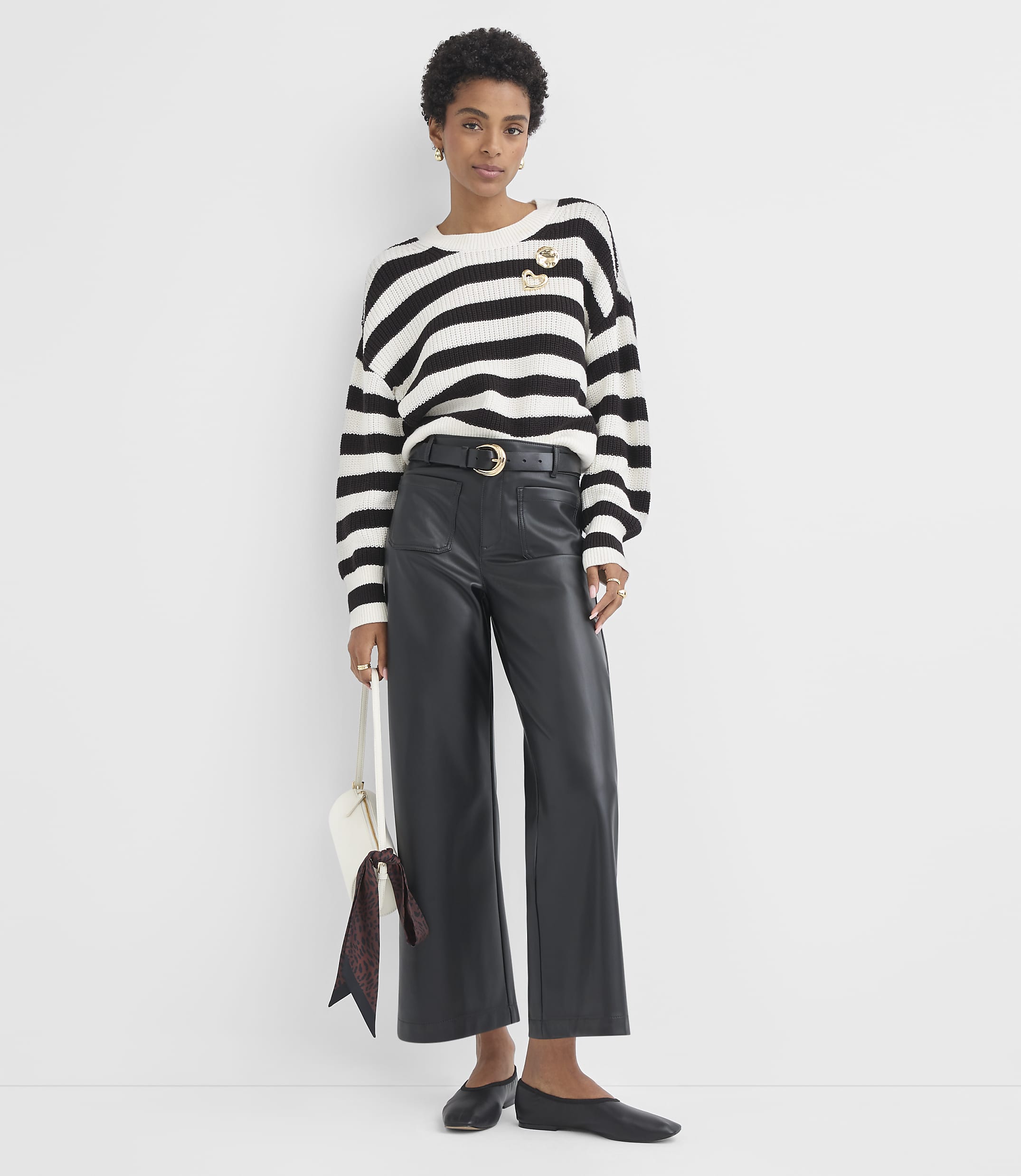 A woman in a striped sweater and leather pants stands against a plain white background, holding a white purse with a patterned scarf.