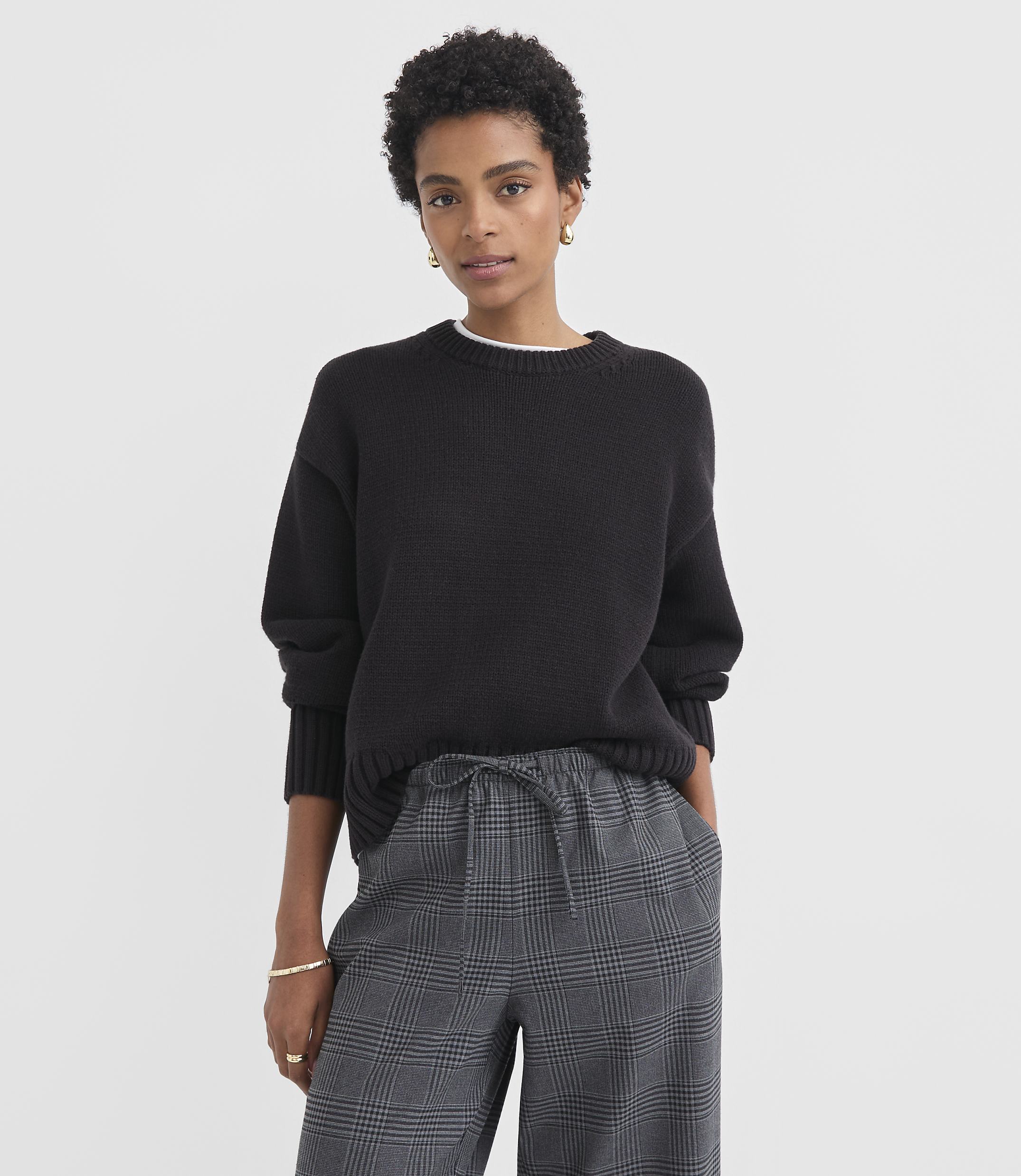 A woman in a black sweater and plaid pants stands against a plain white background.
