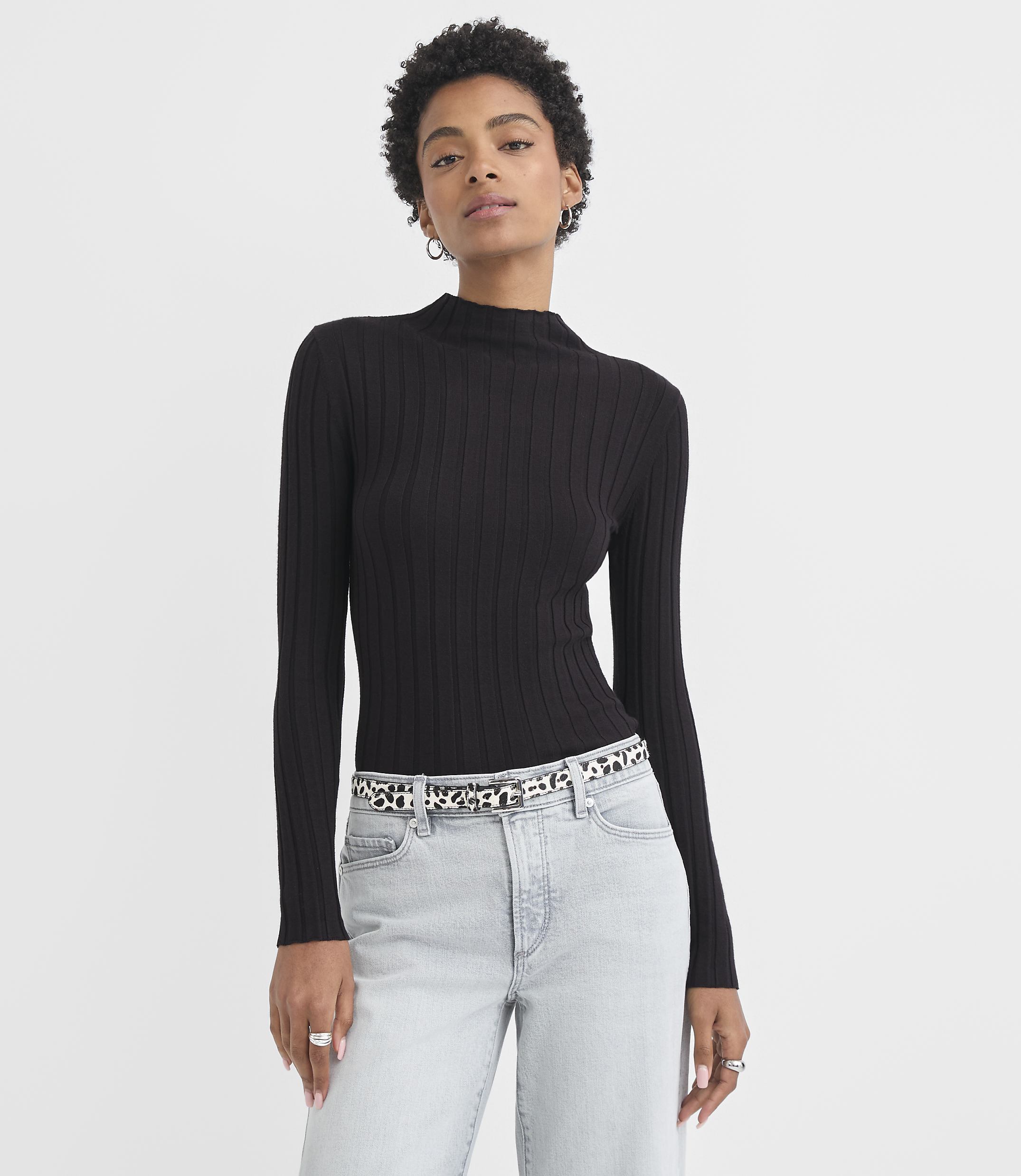 A woman in a black ribbed turtleneck and light wash jeans stands against a plain white background.