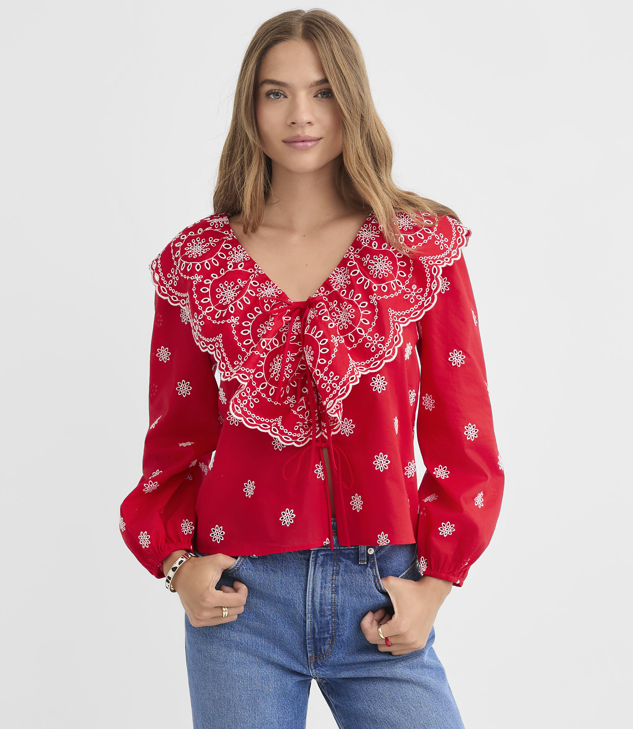 A woman in a red, embroidered blouse and blue jeans stands against a plain white background.