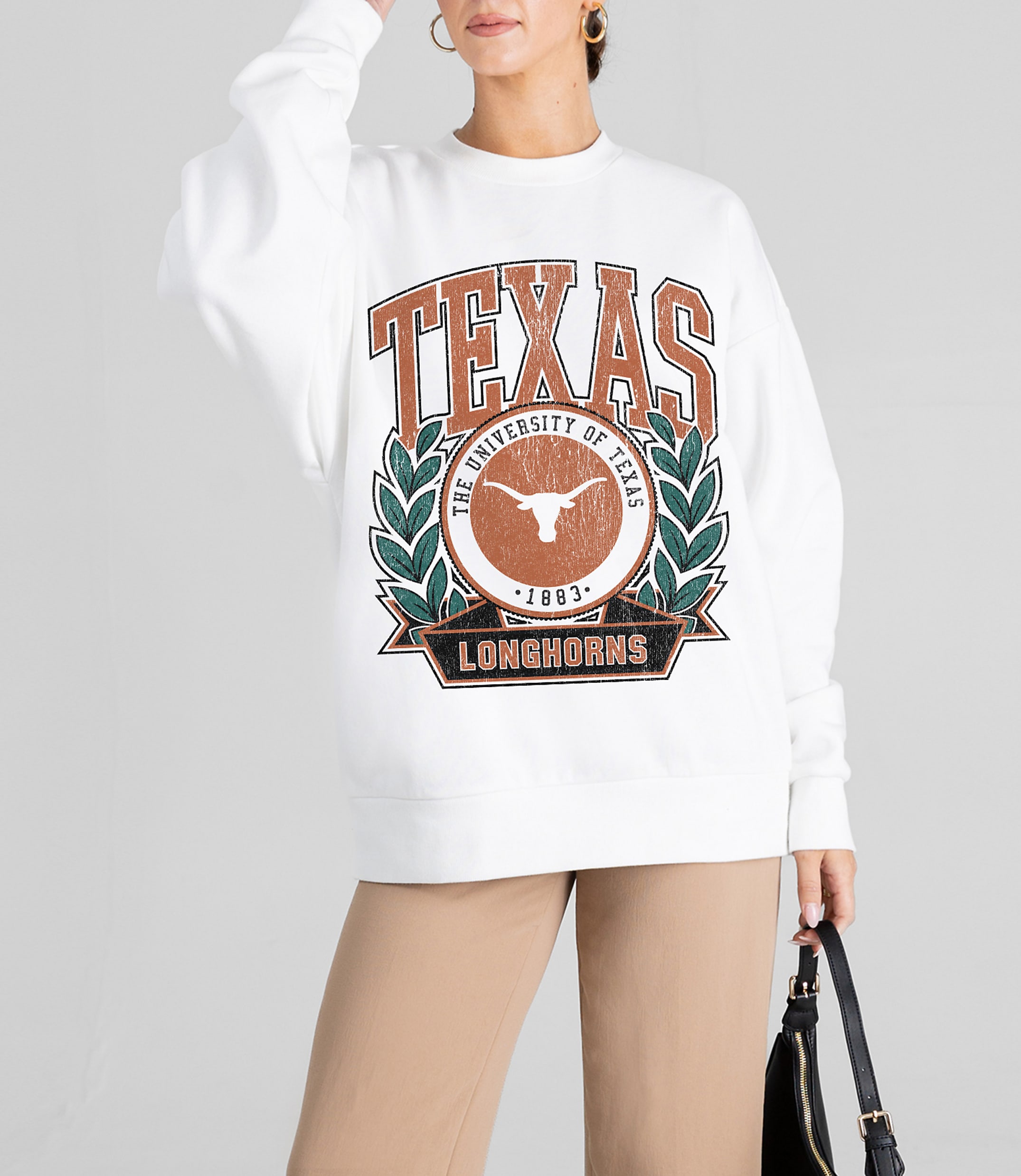 A woman in a white sweatshirt with a University of Texas Longhorns graphic stands against a plain gray background, holding a black purse.